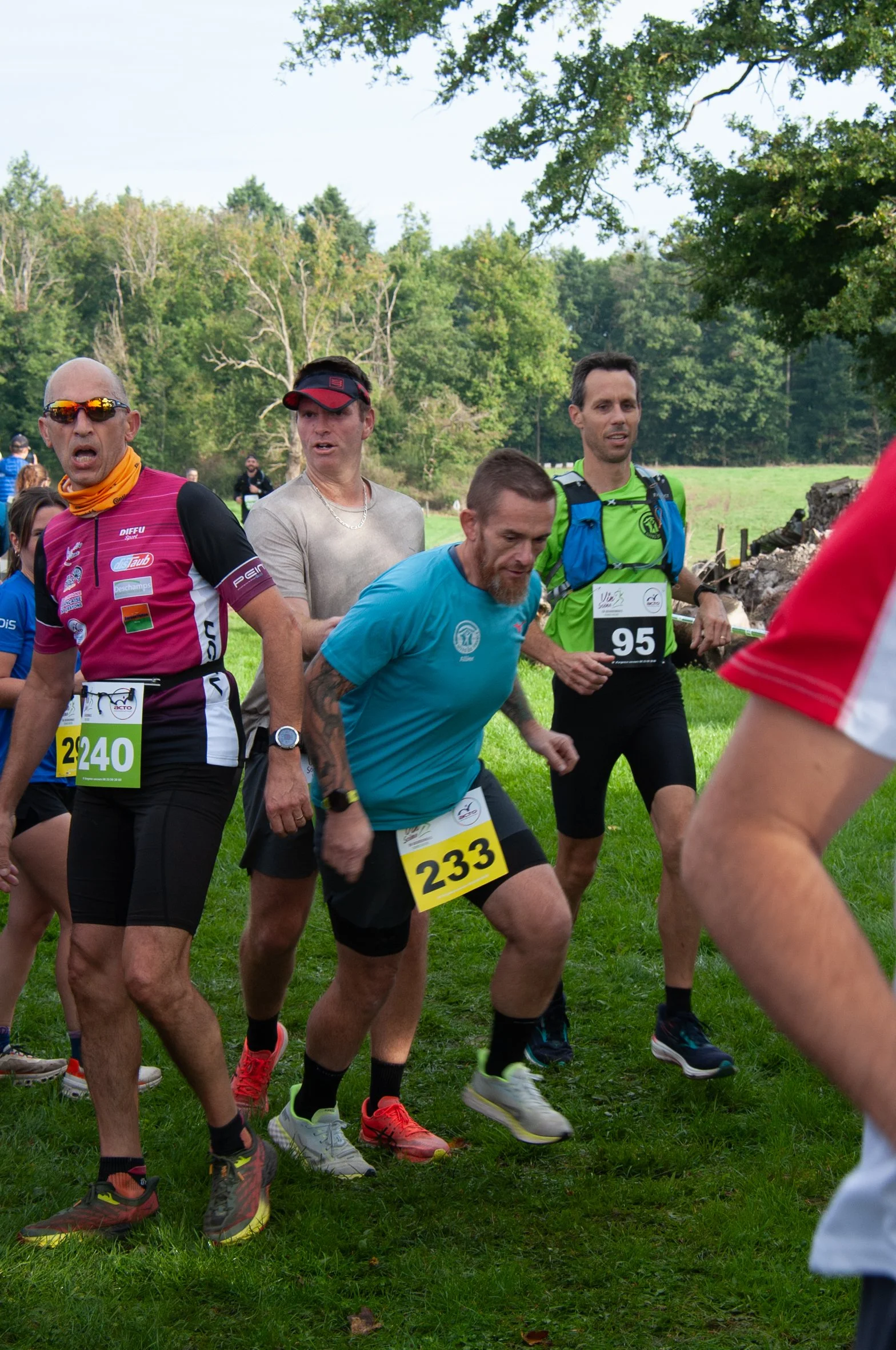Groupe de coureurs participant à une course en plein air, portant des dossards avec des numéros, dans un environnement verdoyant avec des arbres en arrière-plan.