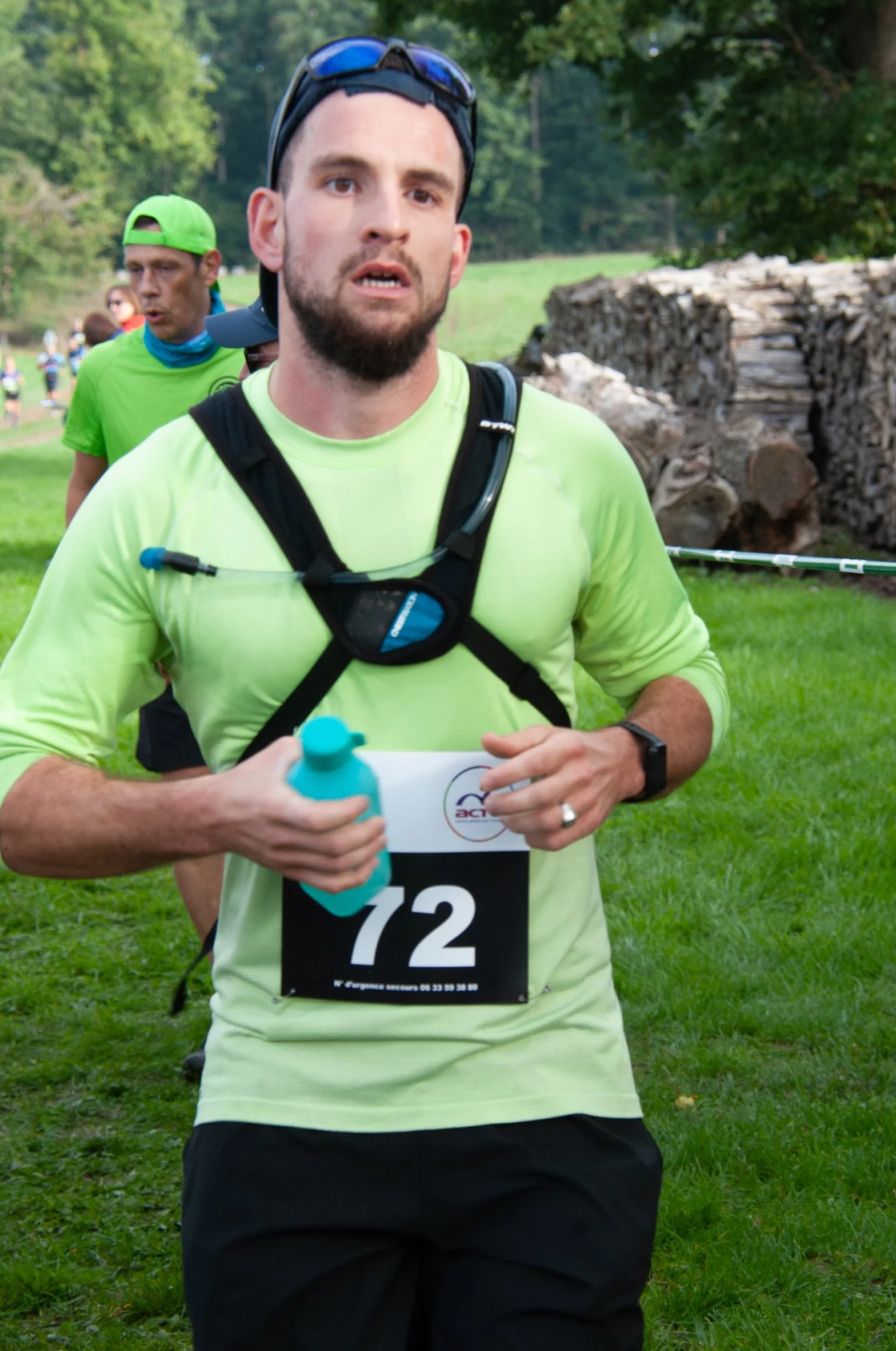 Homme coureur portant un t-shirt vert pâle, portant un gel dans une main, participant à une course dans un parc avec d'autres coureurs en arrière-plan.