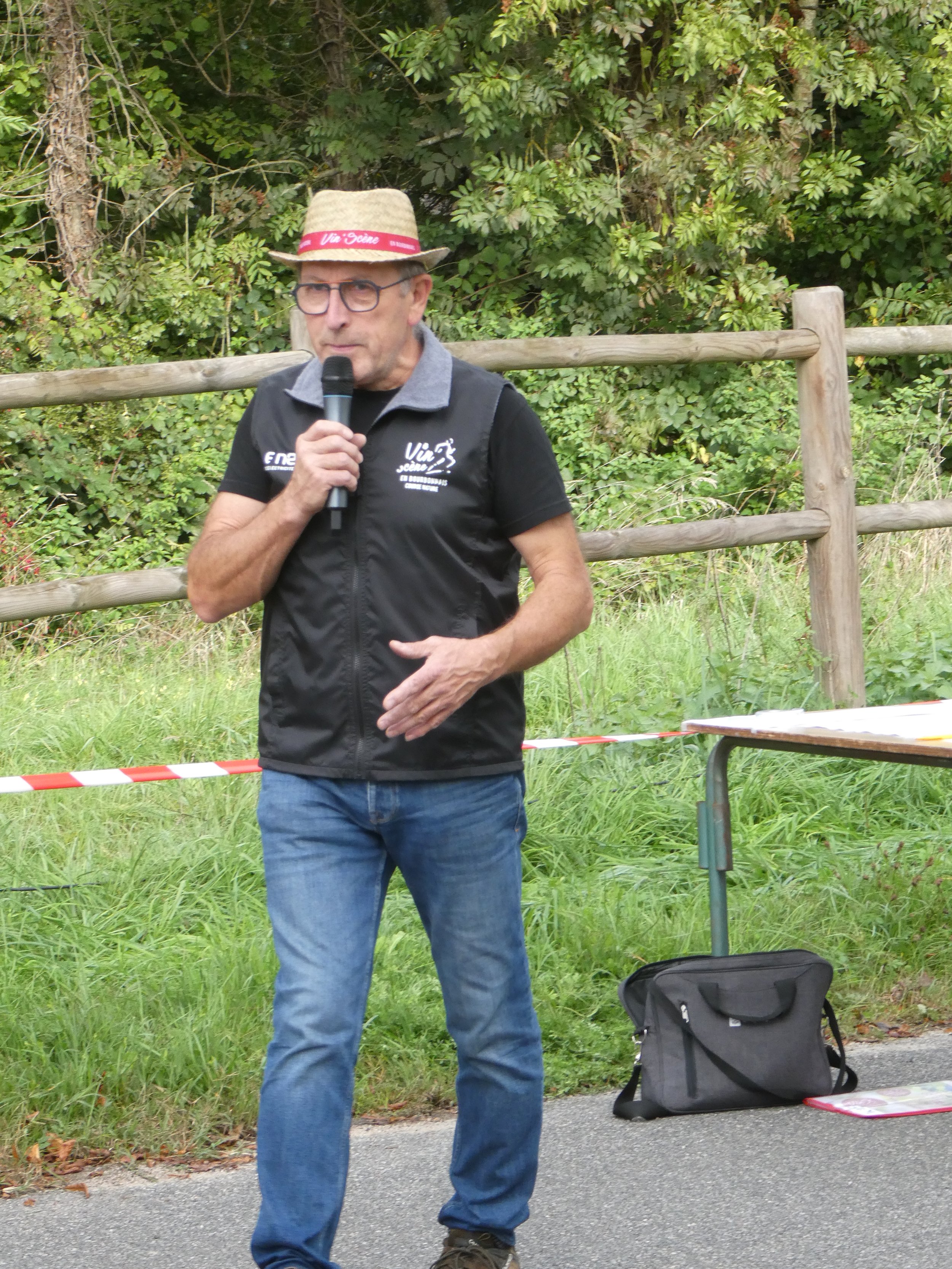 Un homme portant un chapeau, des lunettes et un gilet noir, parlant dans un micro lors d'un événement en extérieur, avec une table et un sac à ses côtés, entouré de verdure.