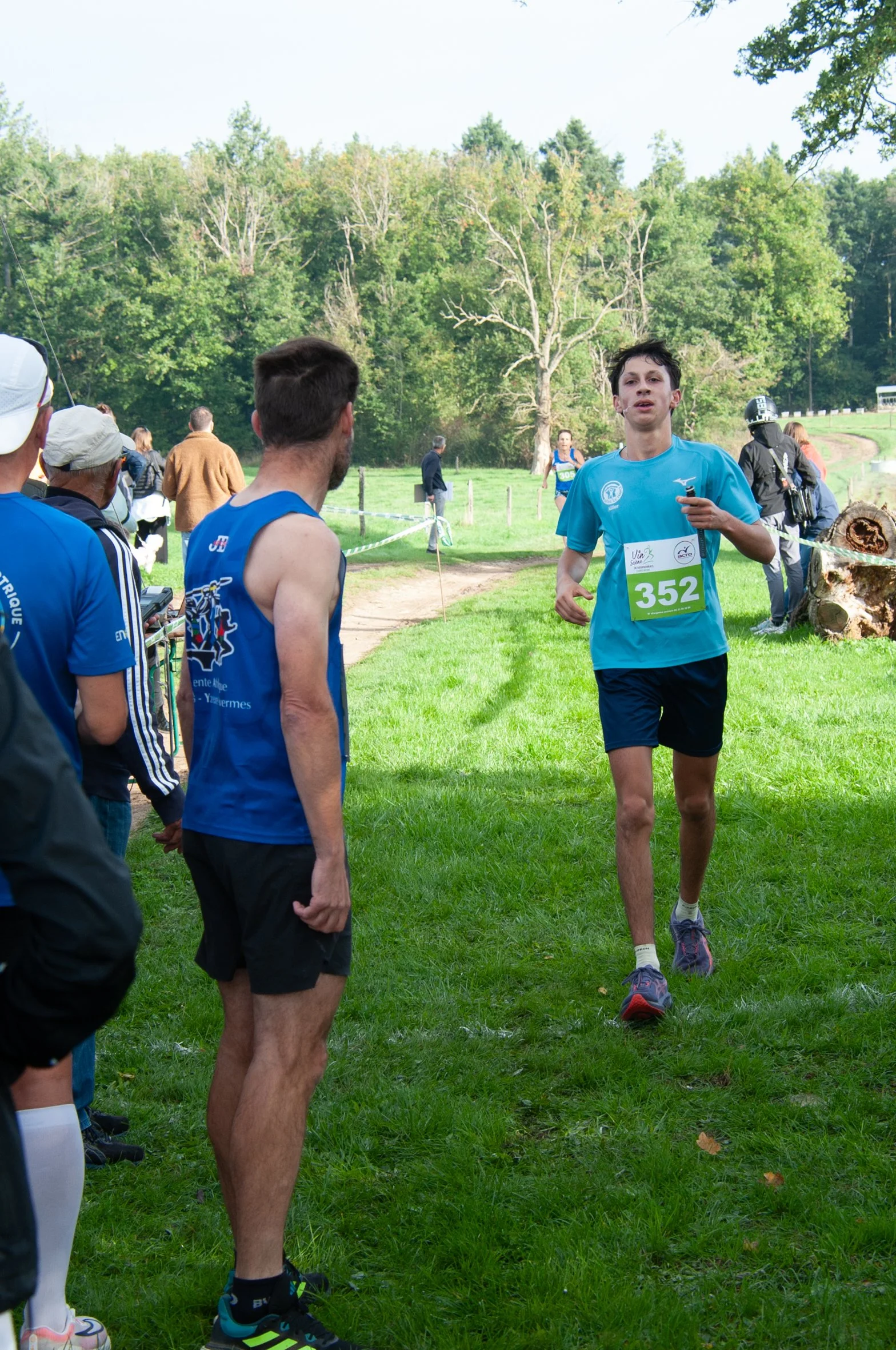 Un jeune coureur traverse la ligne d'arrivée lors d'une course à pied en plein air, entouré de spectateurs sous un ciel clair, avec des arbres verts en arrière-plan.