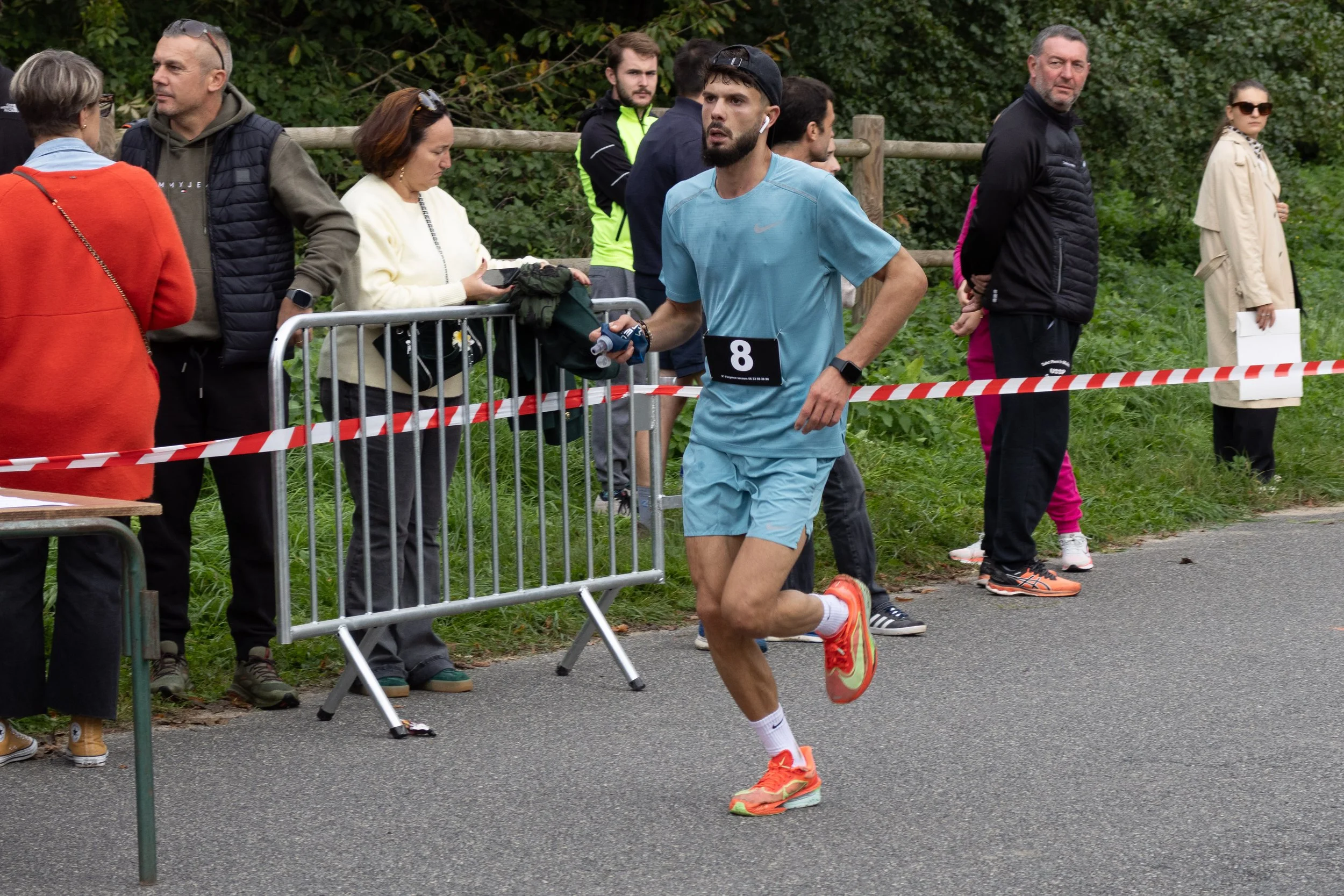 Un homme court lors d'une course, portant un numéro 8, vêtement de sport bleu, chaussures orange et écouteurs, en mouvement sur une route pavée, avec des spectateurs derrière une barrière en arrière-plan.