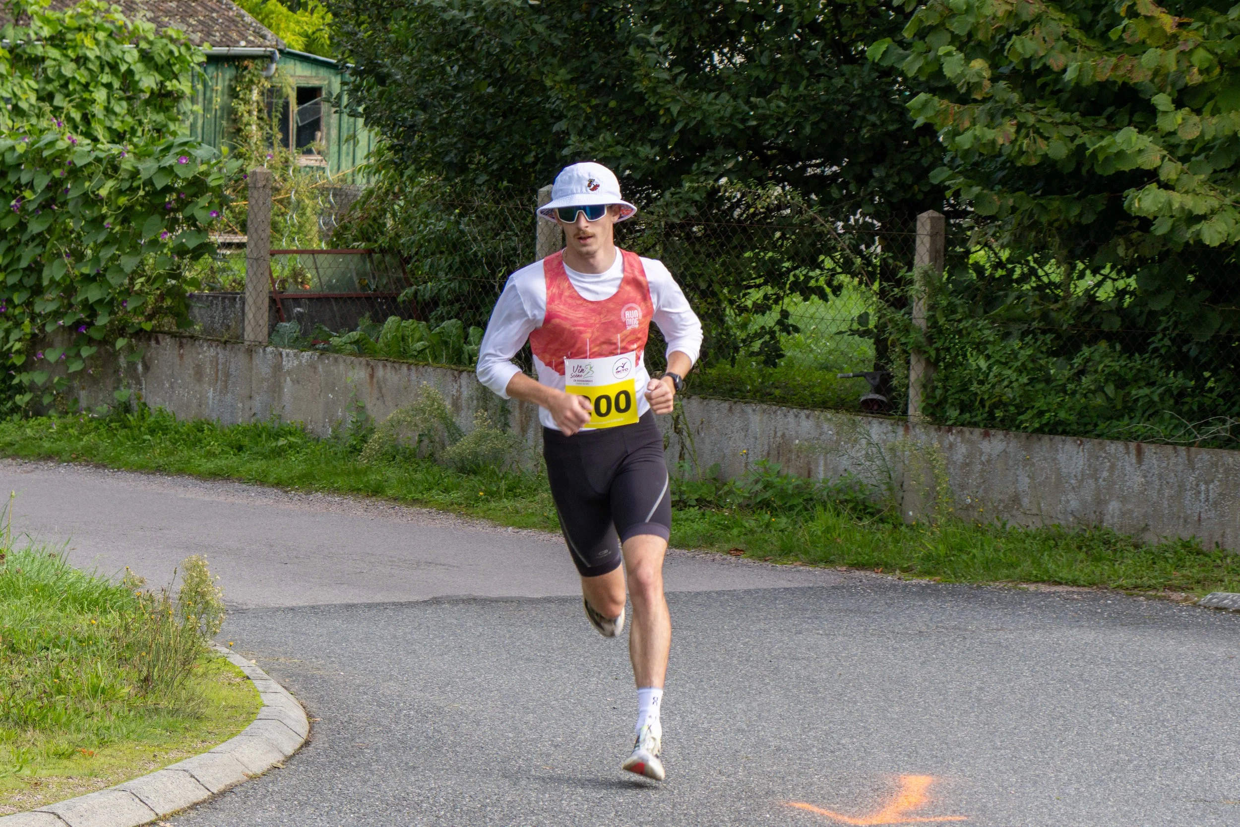 Un jeune homme court sur une route pavée lors d'une course, portant un t-shirt blanc, un gilet rouge, un chapeau blanc, des lunettes de soleil et un dossard avec le numéro 100, entouré de végétation et de clôtures.