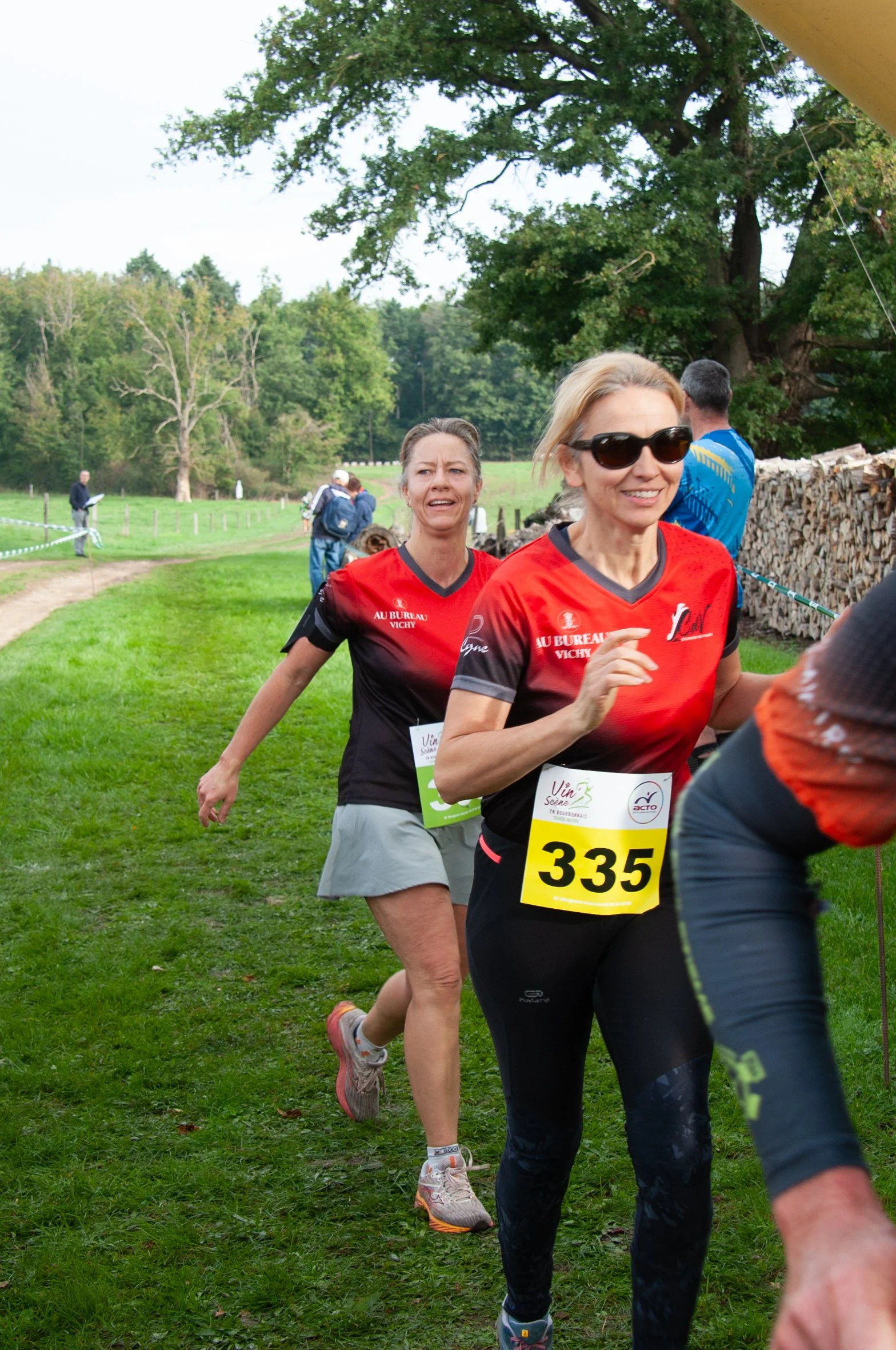 Deux femmes participant à une course en plein air, portant des dossards avec les numéros 335 et d'autres, dans un environnement herbeux avec des arbres et une pile de bois en arrière-plan.