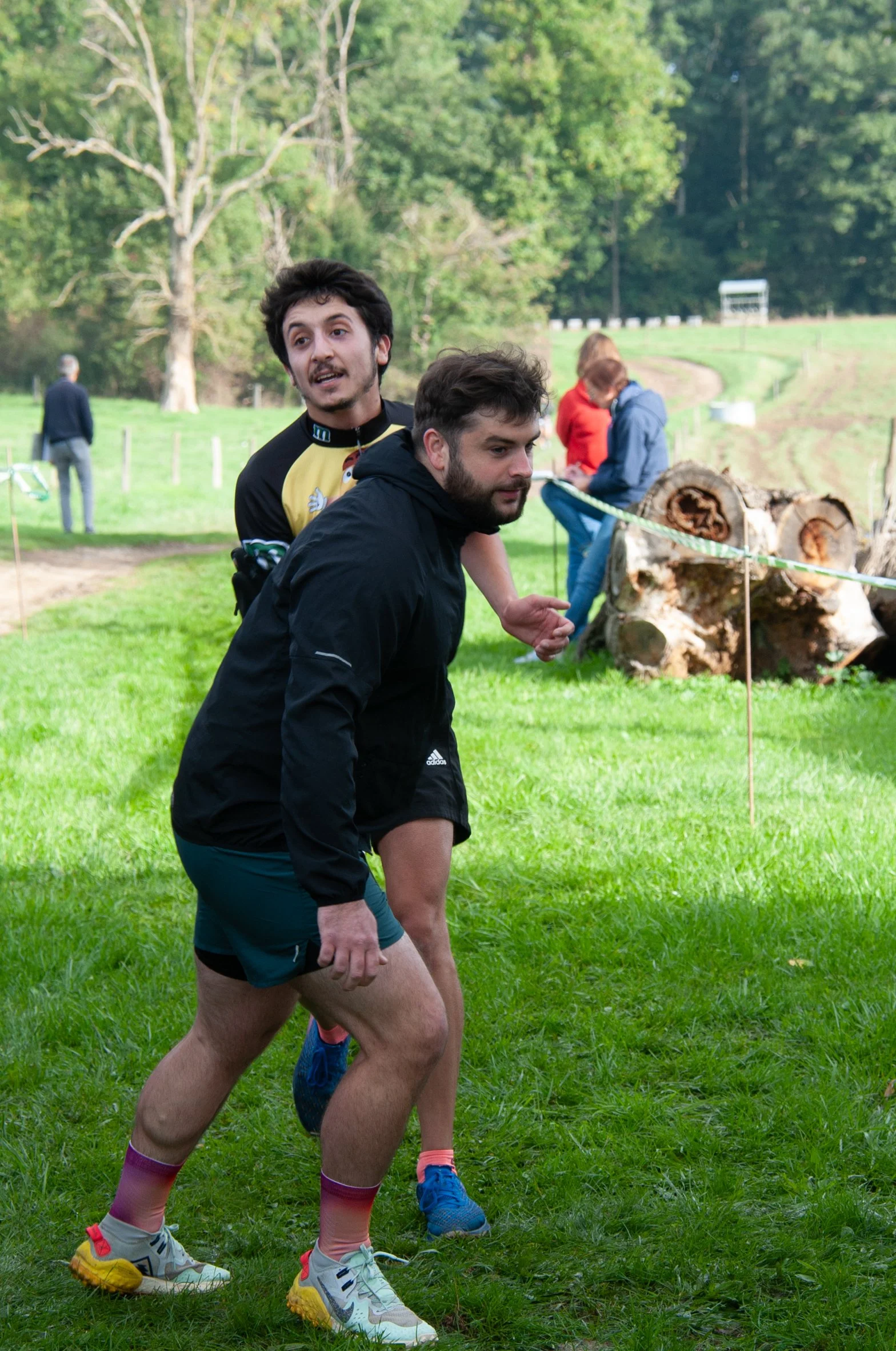 Deux hommes en tenue de sport, en train de courir ou de participer à une activité en plein air sur une pelouse verte, avec des arbres et d'autres personnes en arrière-plan.