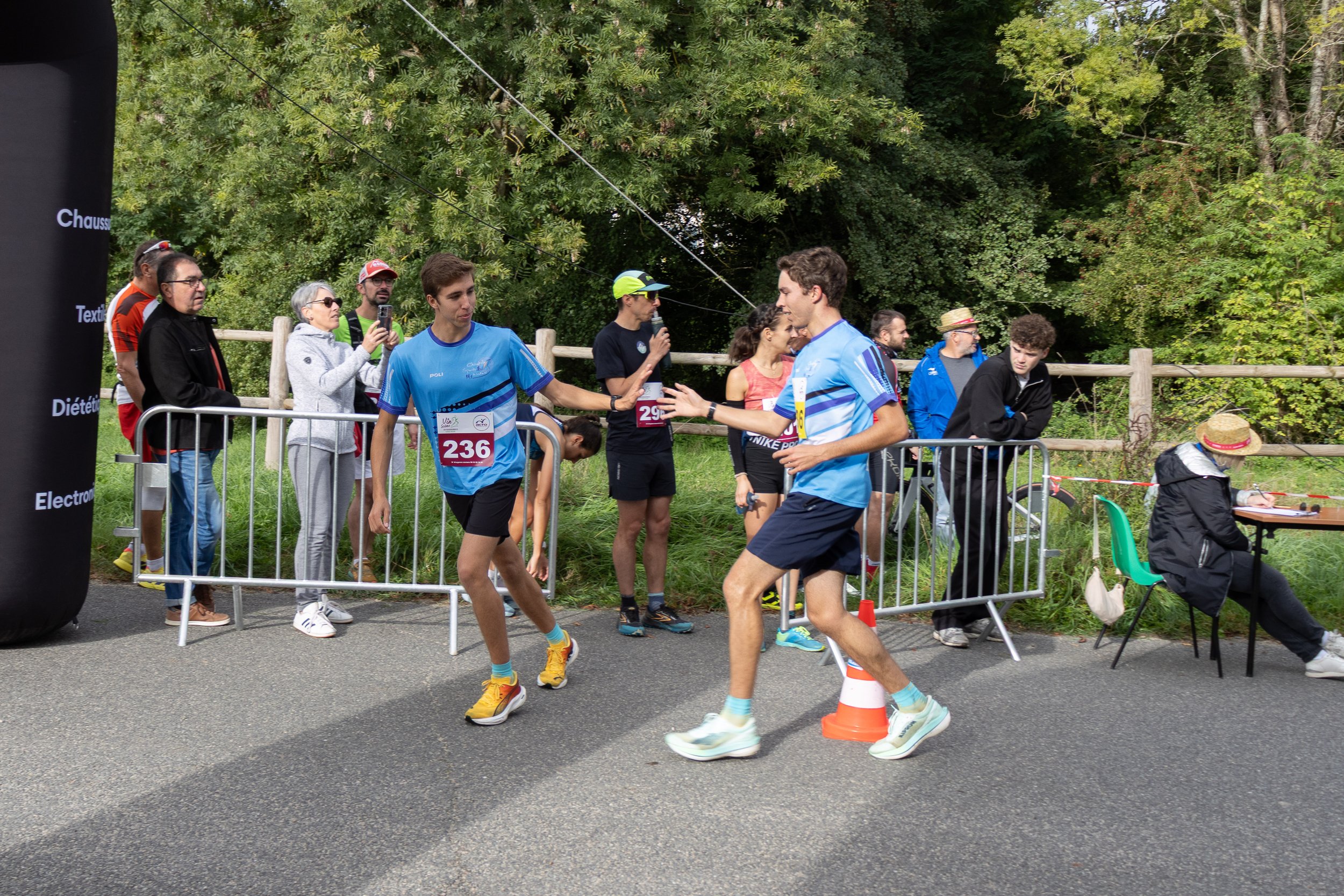 Deux jeunes coureurs en vêtements de course se serrent la main à la fin d'une course, avec des spectateurs et un accueil derrière une barrière.