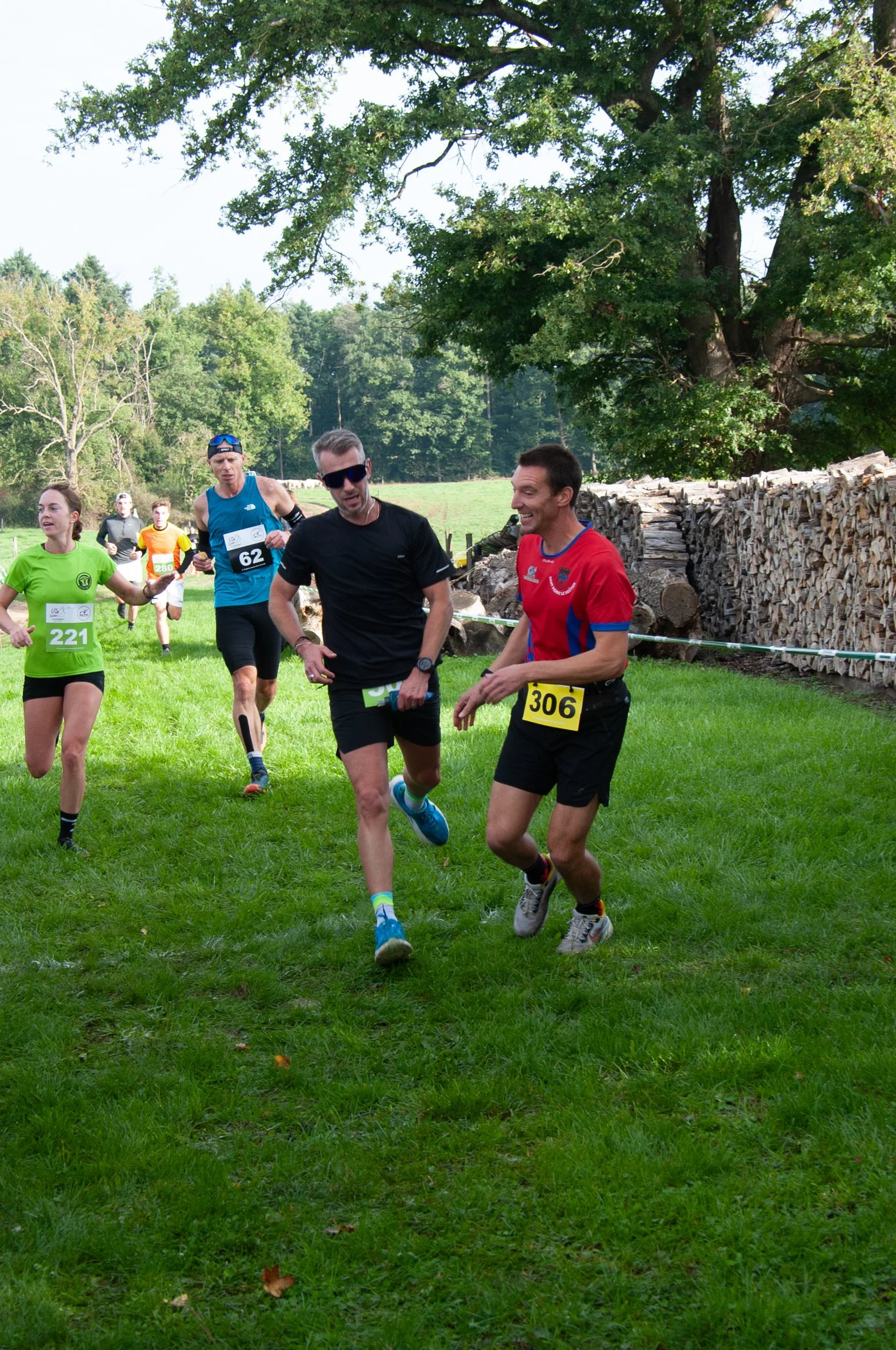 Groupe de coureurs participant à une course en plein air, sur une pelouse verte, avec des arbres en arrière-plan et des tas de bois empilés.