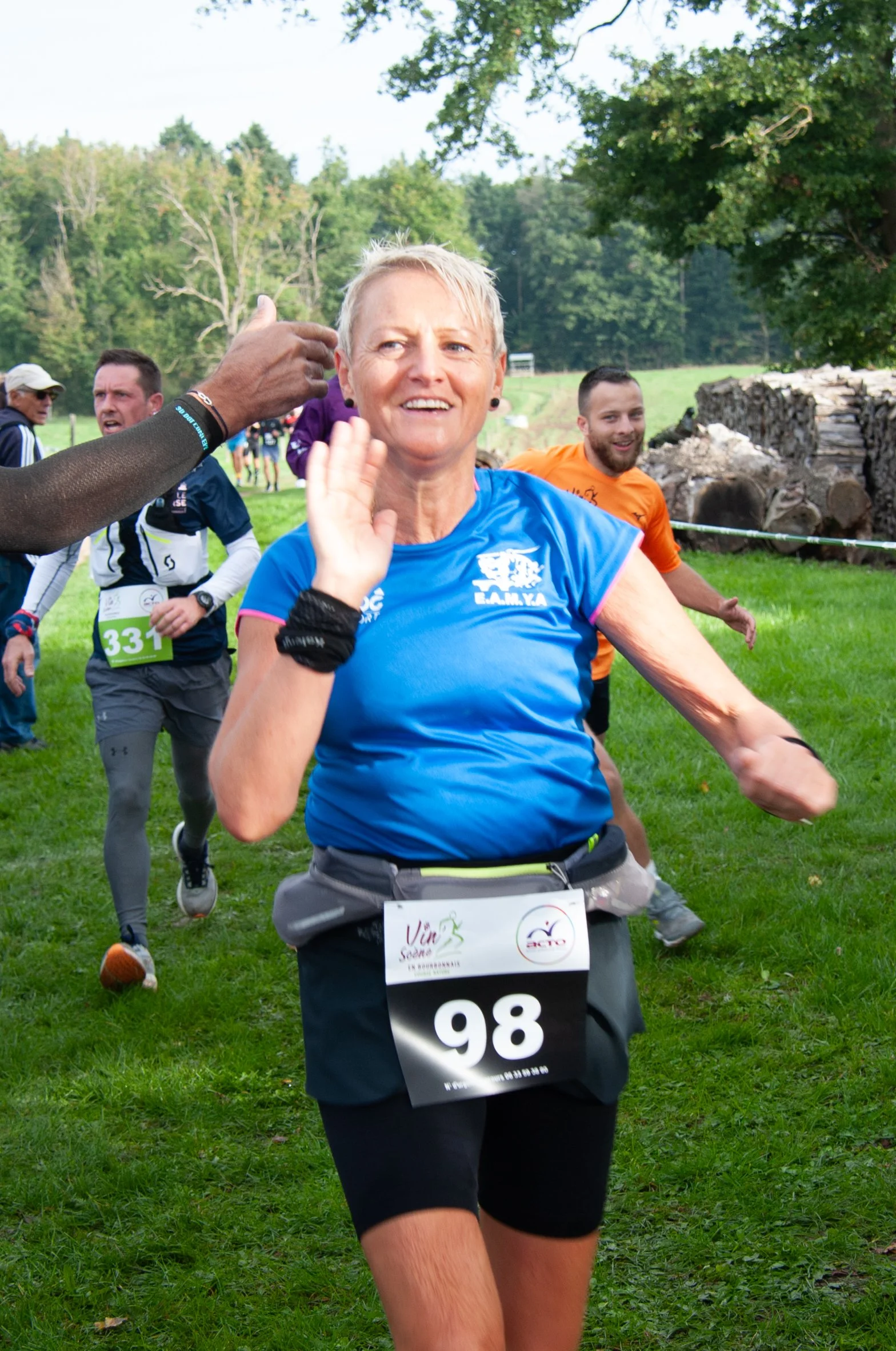 Une femme courante dans une course en extérieur, portant un t-shirt bleu, un numéro 98, et des vêtements de sport, avec d'autres participants en arrière-plan dans un environnement vert avec des arbres.