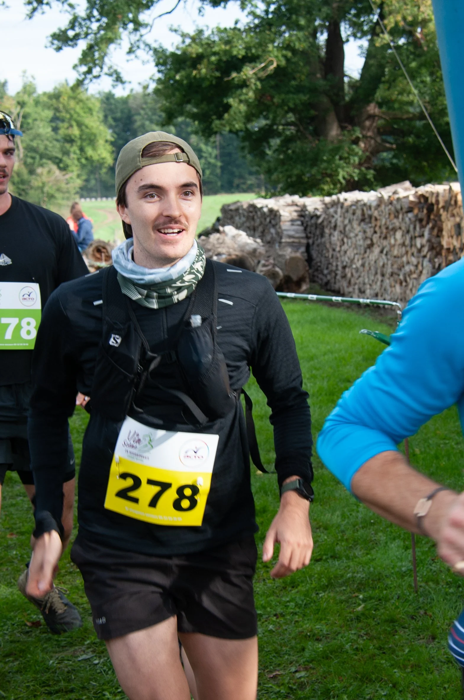 Un homme souriant portant un maillot de course avec le numéro 278, une casquette, et un foulard, en plein marathon dans un environnement verdoyant.