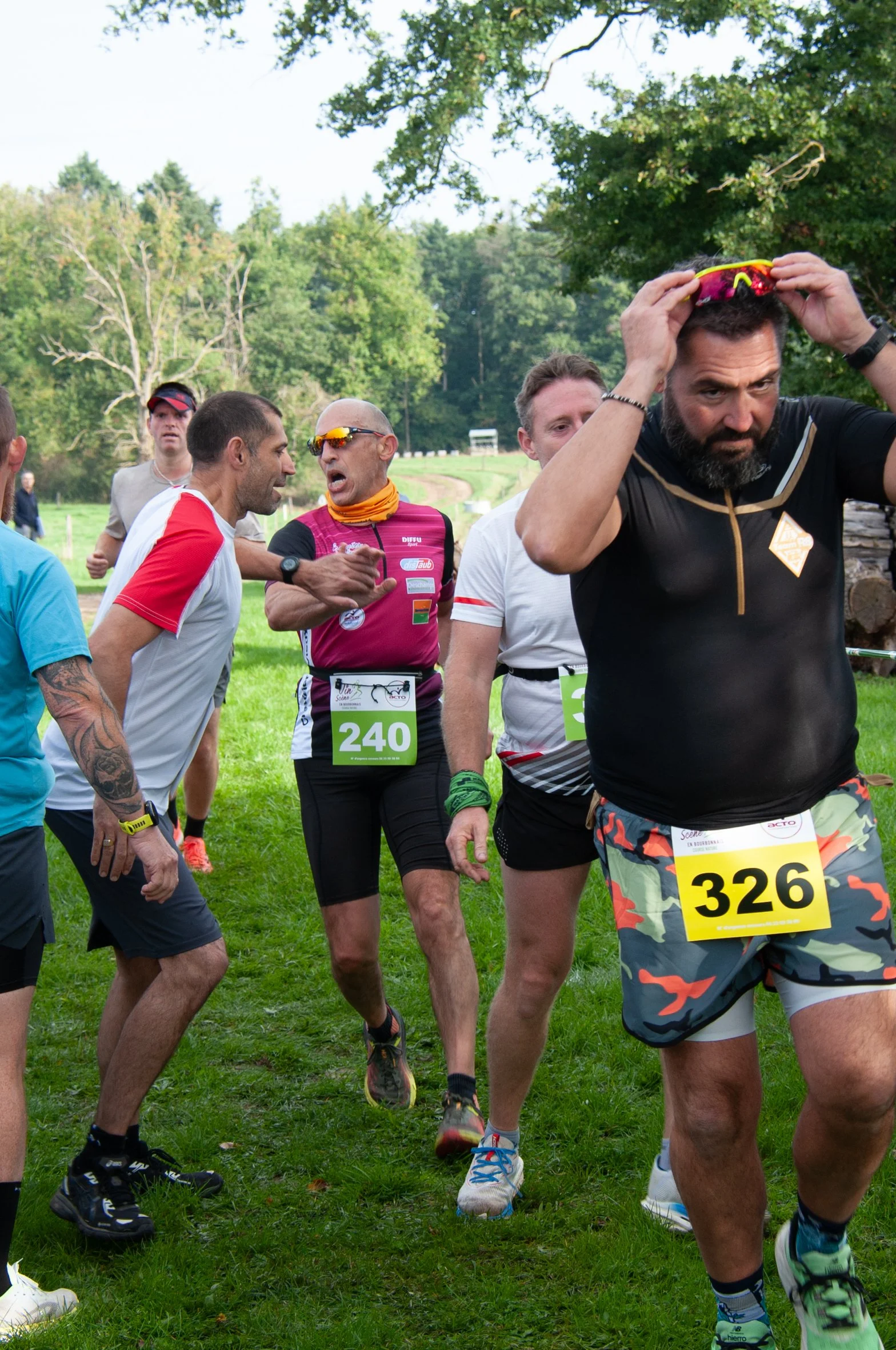Groupe de coureurs de marathon avant la course dans un parc vert, certains discutent et ajustent leur équipement, portant des dossards numérotés.