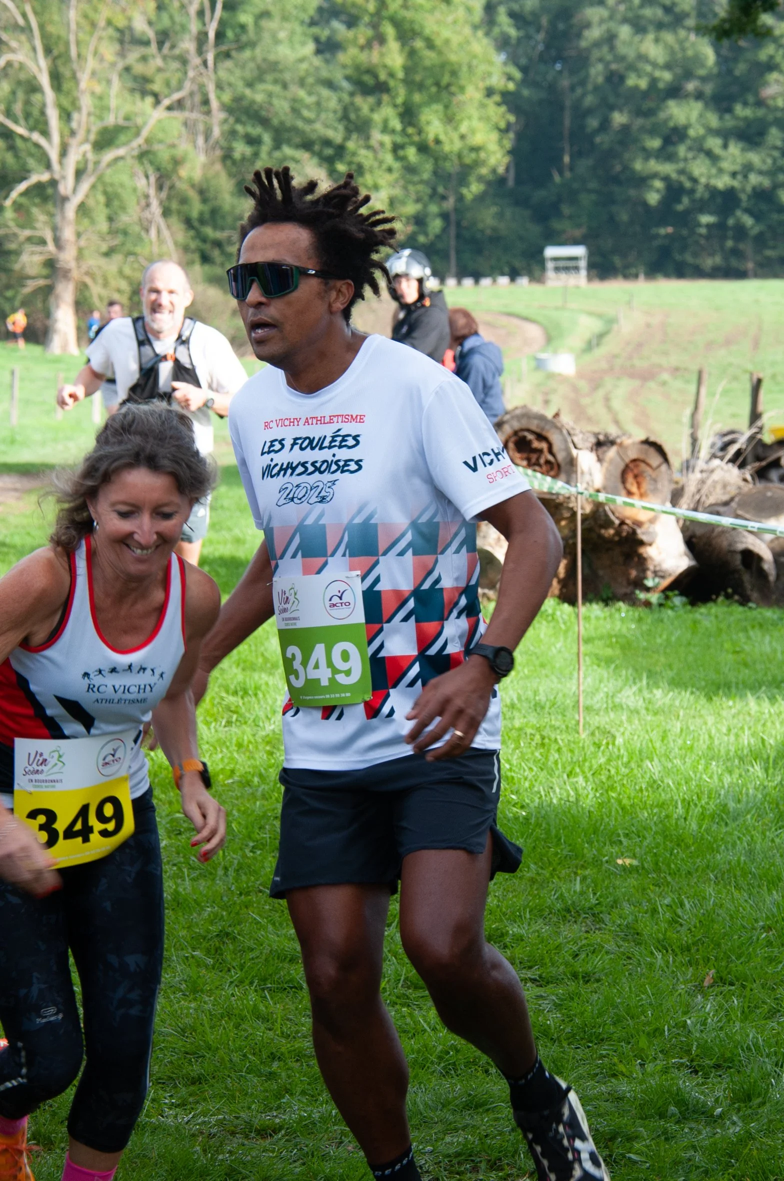 Deux coureurs, un homme et une femme, participants à une course, courent dans un espace vert avec des arbres et un tas de bûches en arrière-plan.