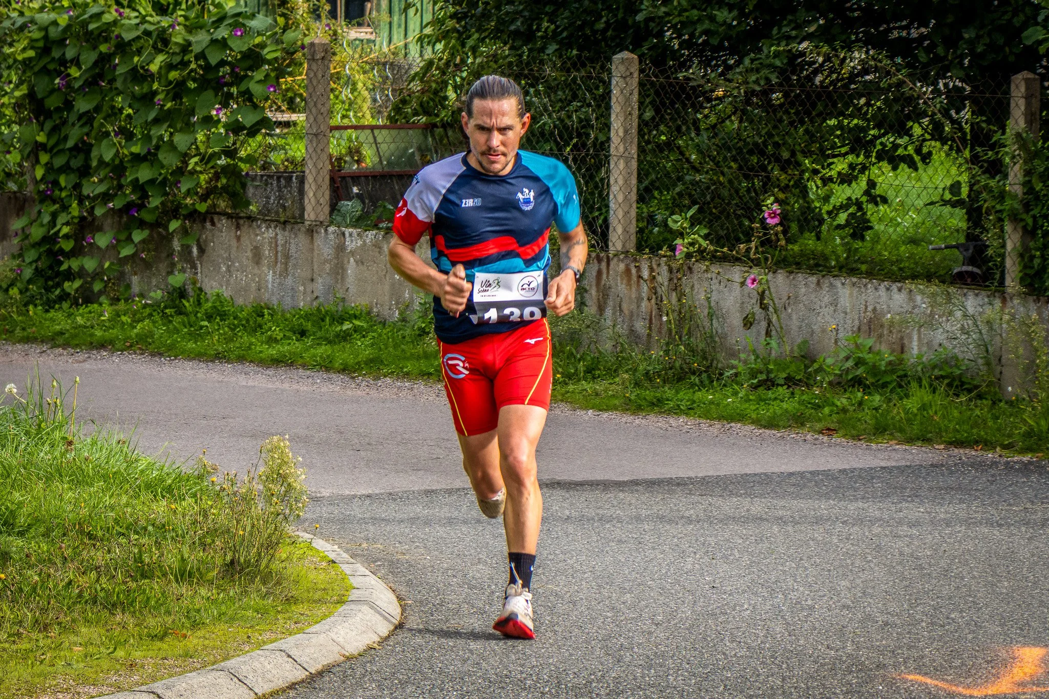 Un homme court sur une route en dehors d'un environnement urbain, portant une tenue de course rouge et bleue avec un numéro 120, entouré de végétation et d'une clôture en arrière-plan.