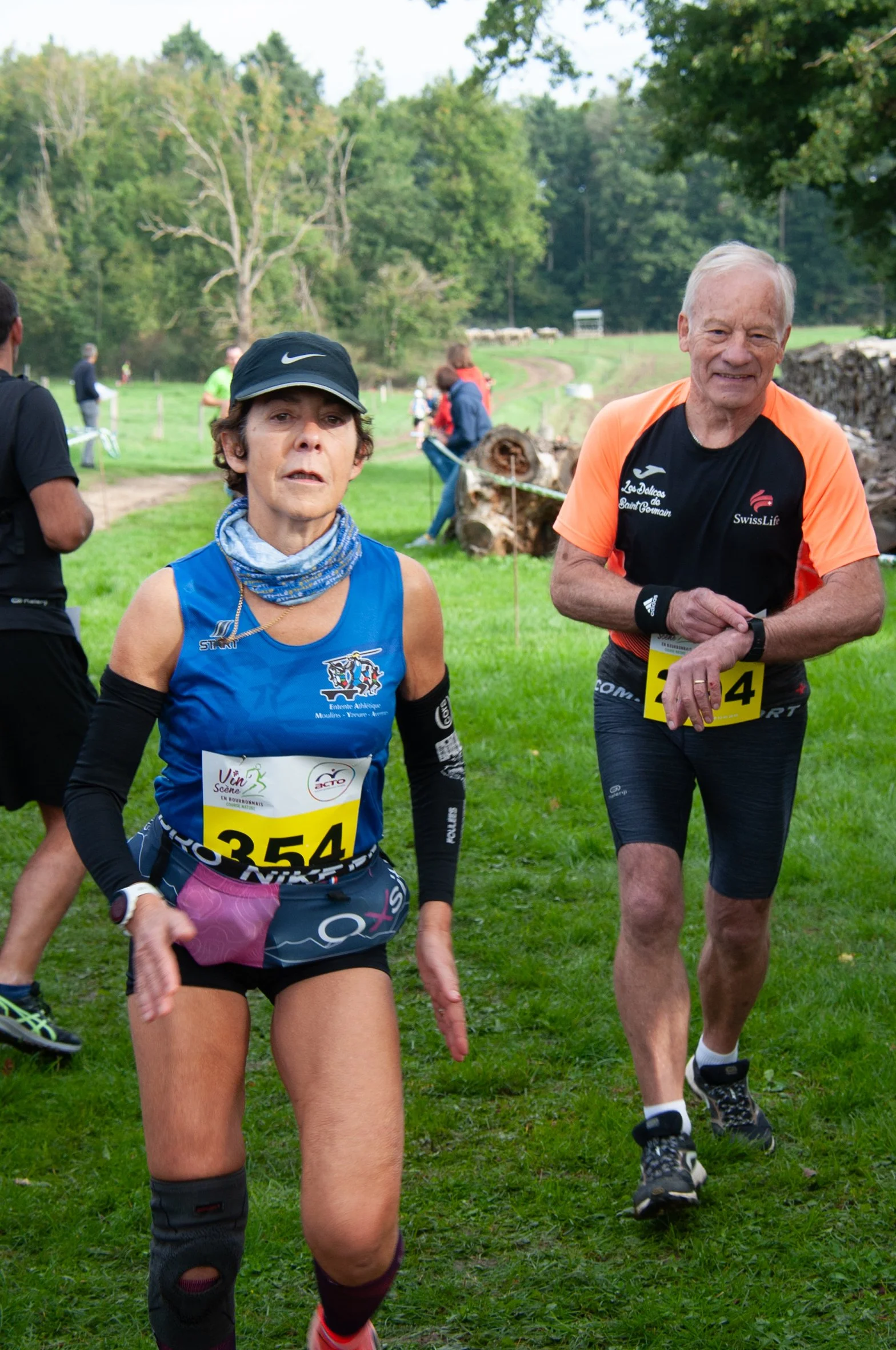 Deux coureurs participent à une course en plein air dans un environnement verdoyant, avec d'autres participants en arrière-plan.