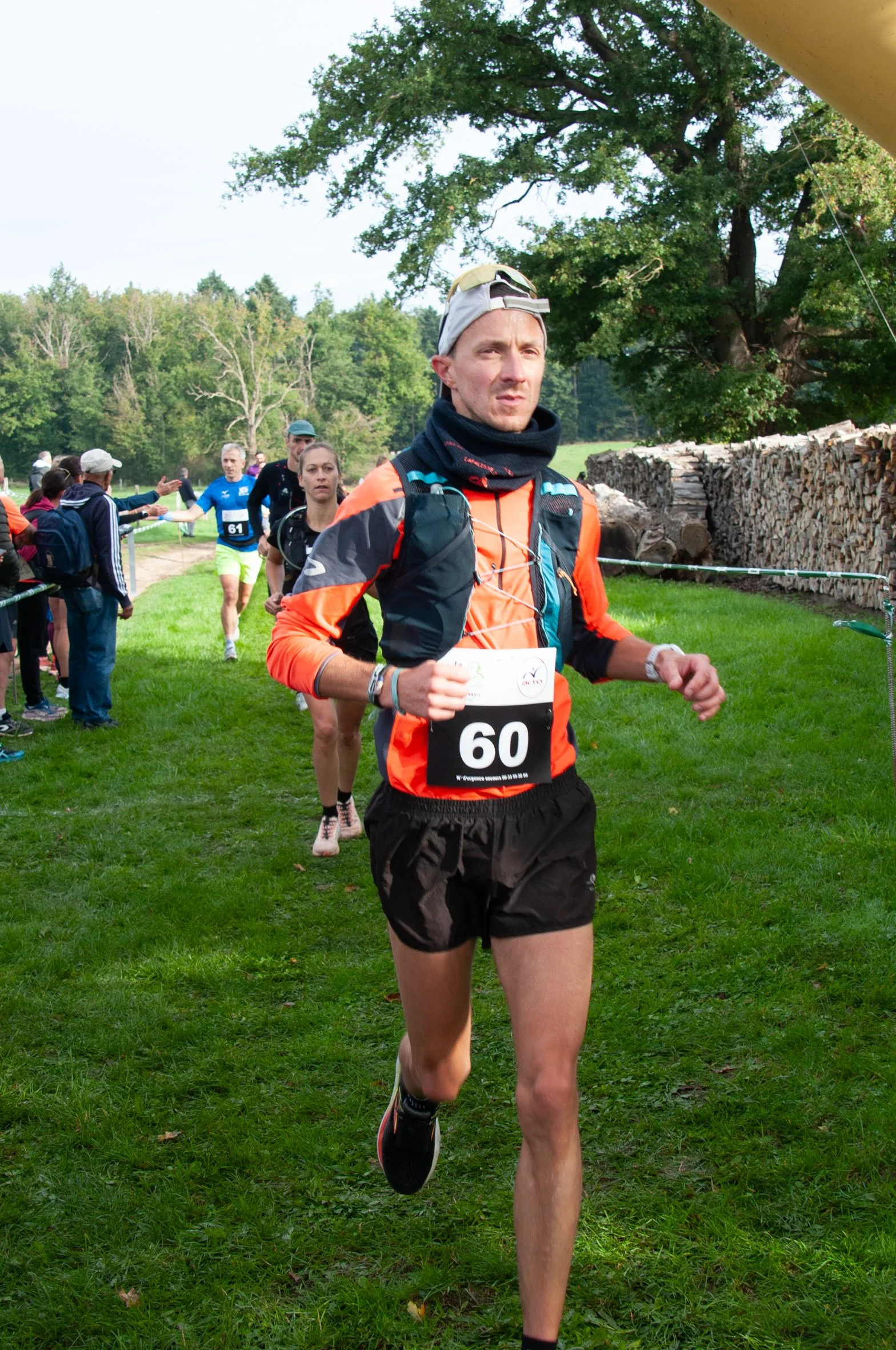 Un coureur portant un maillot avec le numéro 60 participe à une course en extérieur, entouré d'autres coureurs et spectateurs dans un environnement naturel avec arbres et bois.