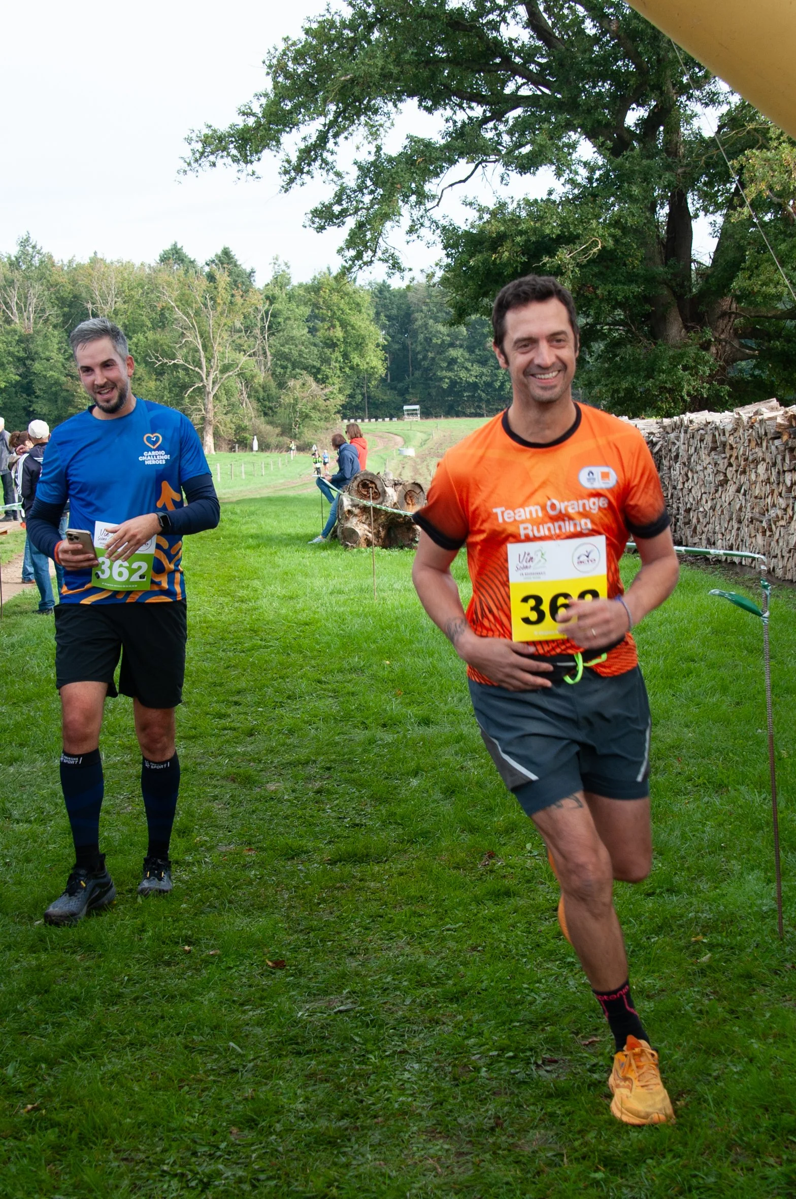 Deux hommes courent lors d'une course en plein air dans une zone verte avec des arbres. La personne en avant porte un t-shirt orange avec le texte 'Team Orange Running' et un dossard numéro 369. La seconde personne porte un t-shirt bleu avec le numér