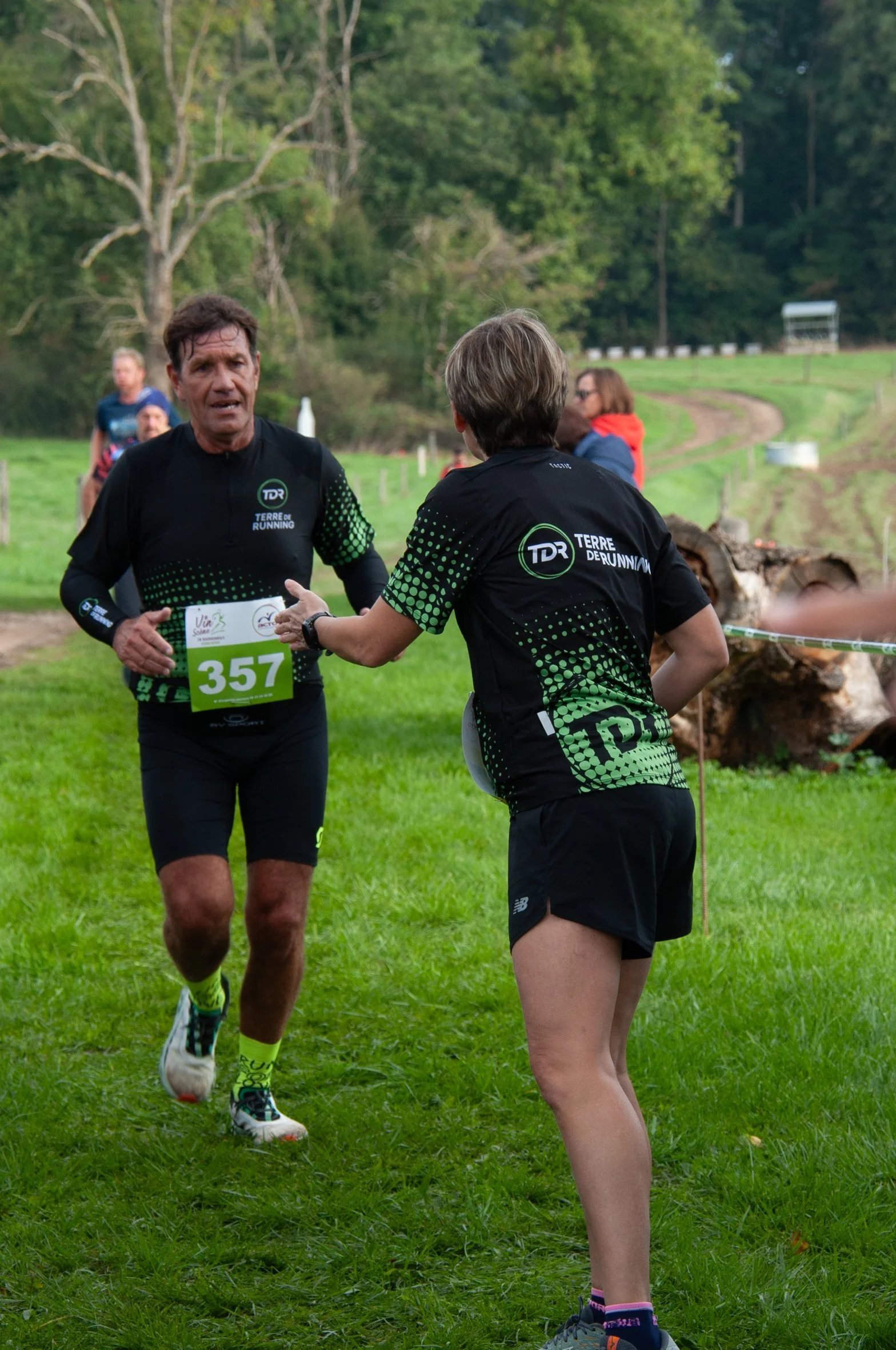 Deux coureurs en pleine course d'un marathon, l'un donne quelque chose à l'autre en dehors du parcours, dans un environnement vert avec des arbres et une pelouse.