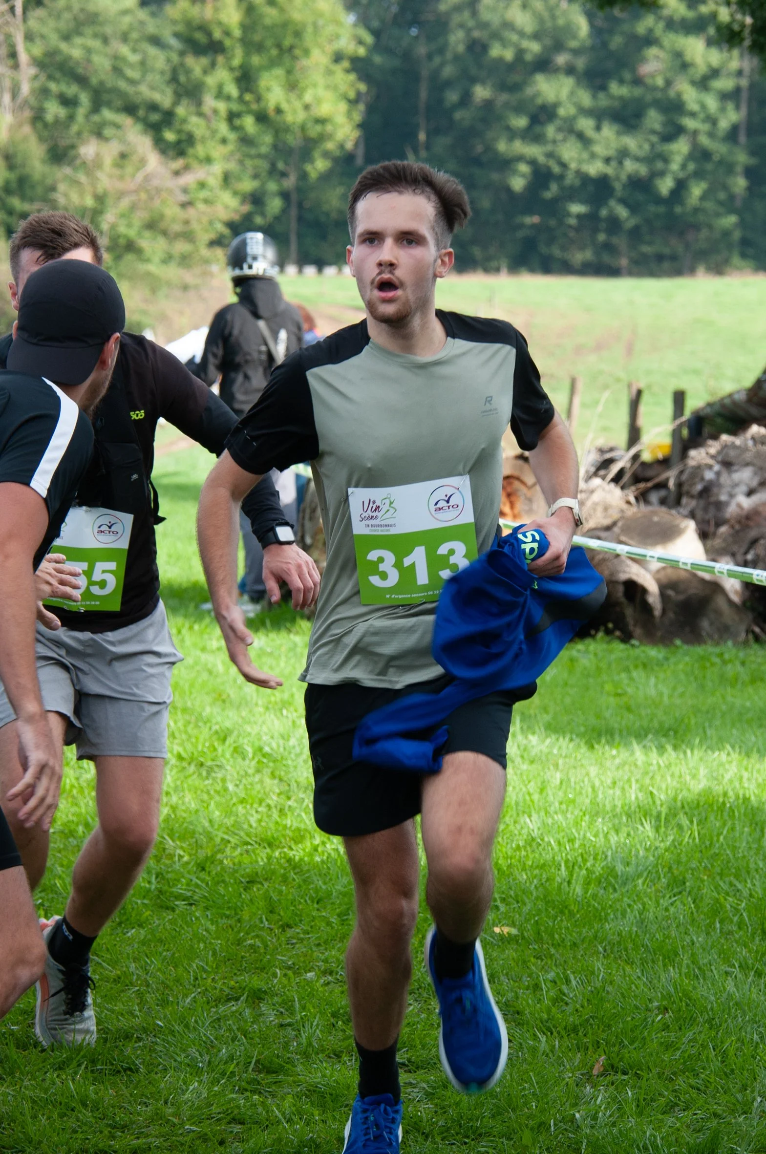 Un homme courant lors d'une course en plein air, portant un numéro 313, avec plusieurs autres participants autour, dans un paysage verdoyant.