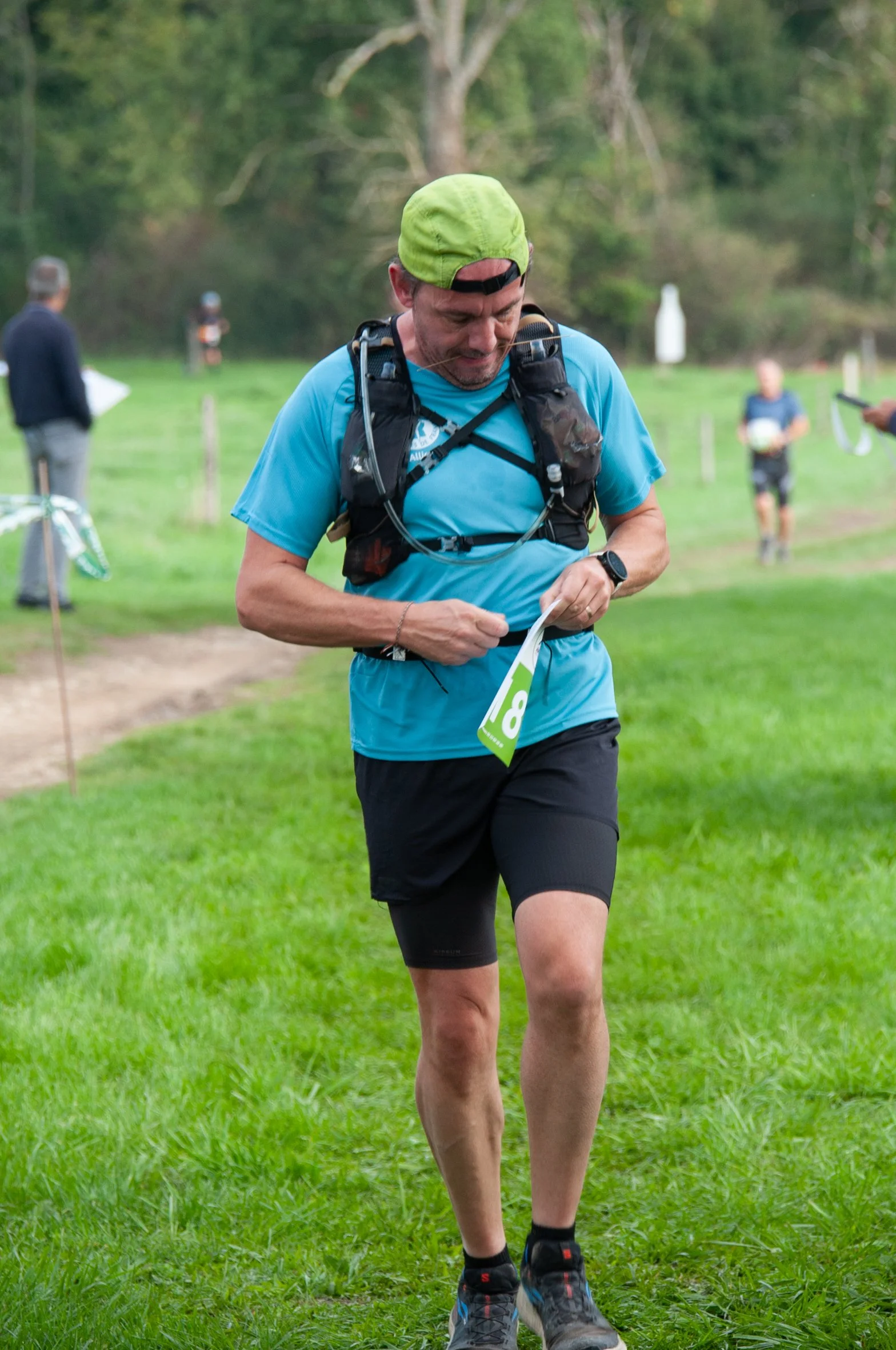 Homme courant dans un parc verdoyant avec un sac à dos, portant un t-shirt bleu et un casque, en pleine activité de trail ou course nature.