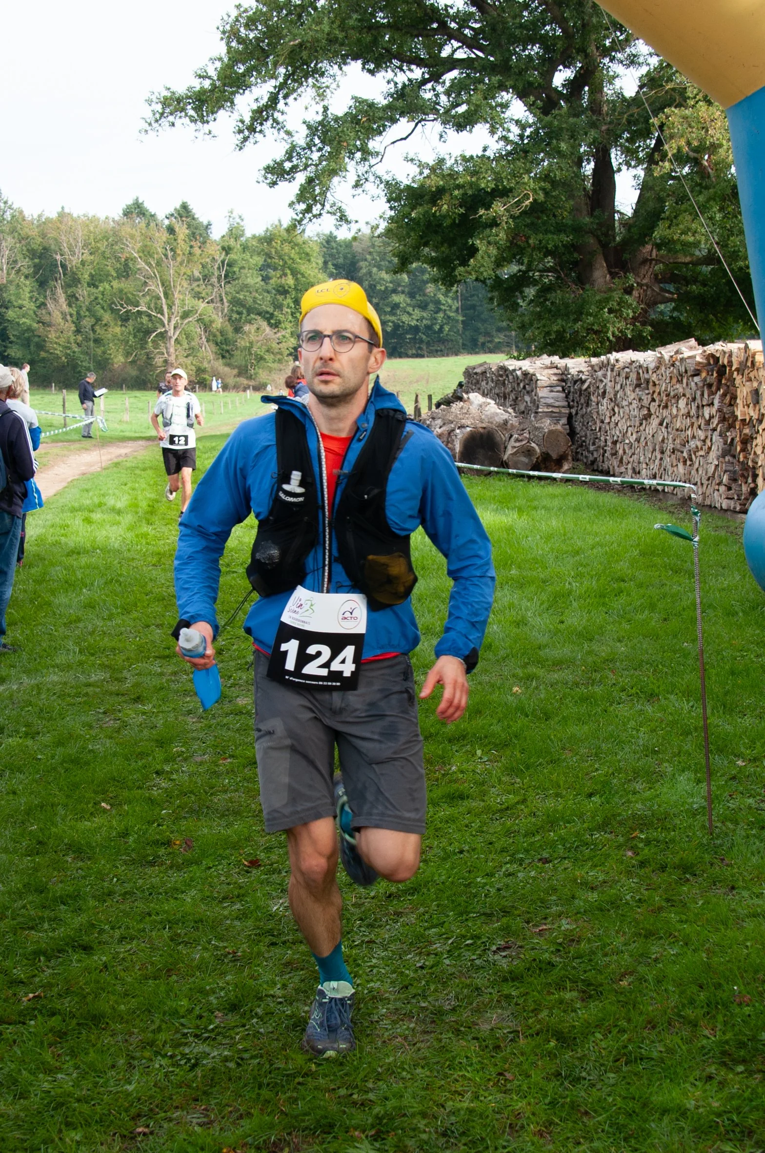 Un homme court lors d'une course en plein air, portant un maillot avec le numéro 124, une veste bleue, un chapeau jaune, et des lunettes.