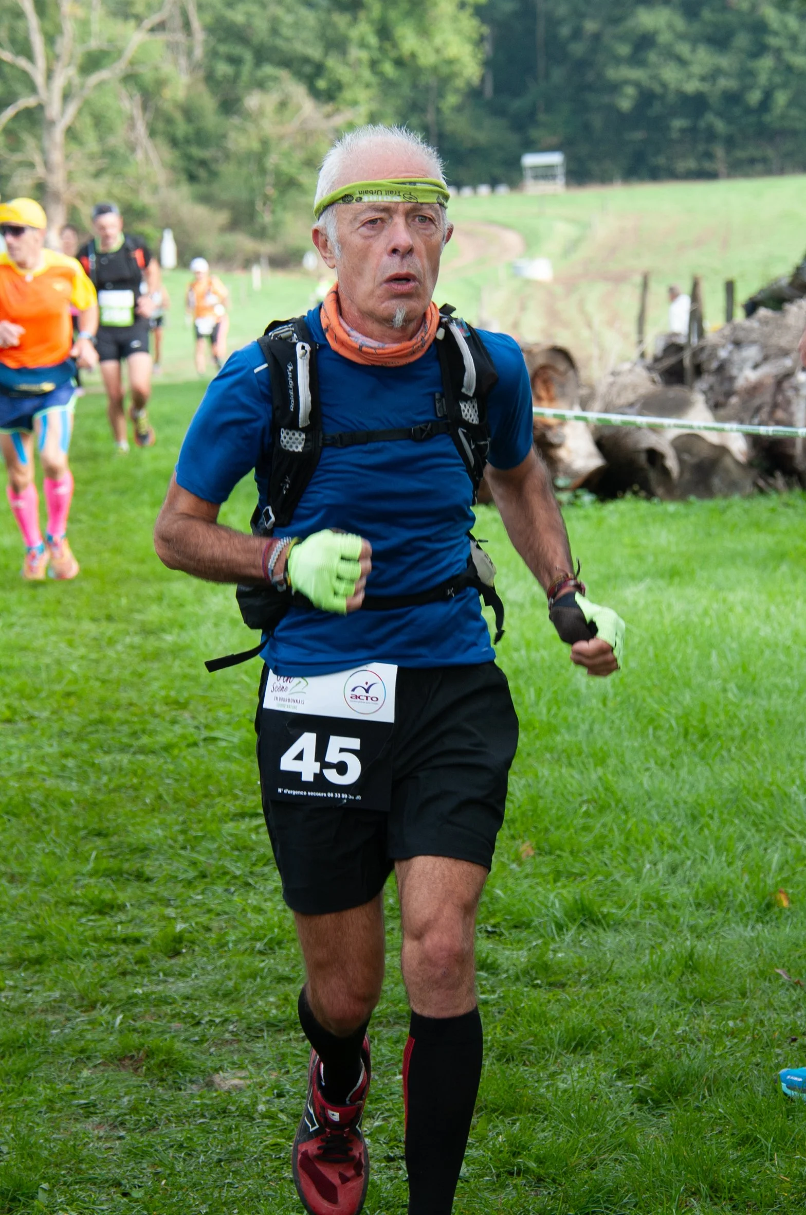 Un homme âgé court lors d'une course en plein air, portant un t-shirt bleu, un casque de course, un sac à dos, des gants verts, un numéro 45, et des vêtements de sport. D'autres coureurs sont visibles en arrière-plan.