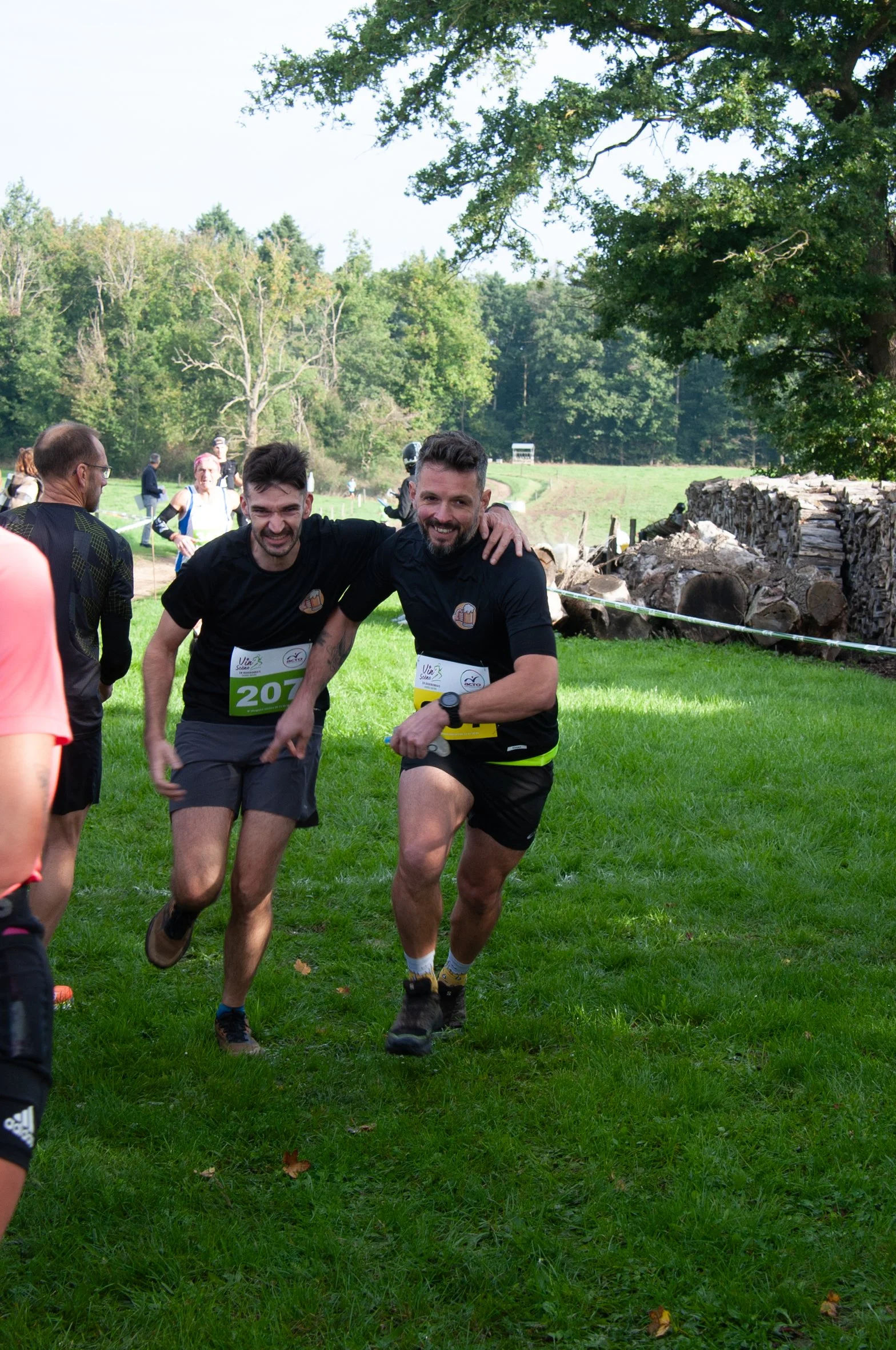 Deux hommes souriants participent à une course en plein air, courant sur une pelouse verte avec des arbres en arrière-plan.