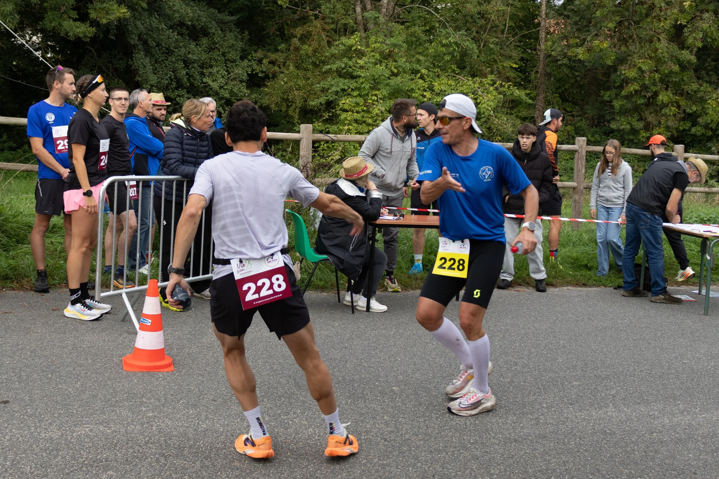 Deux coureurs en tenue de course partageant un moment de célébration ou de discussion commerciale près de la ligne d'arrivée lors d'une compétition, avec un groupe de spectateurs et organisateurs en arrière-plan dans un environnement extérieur verdoy