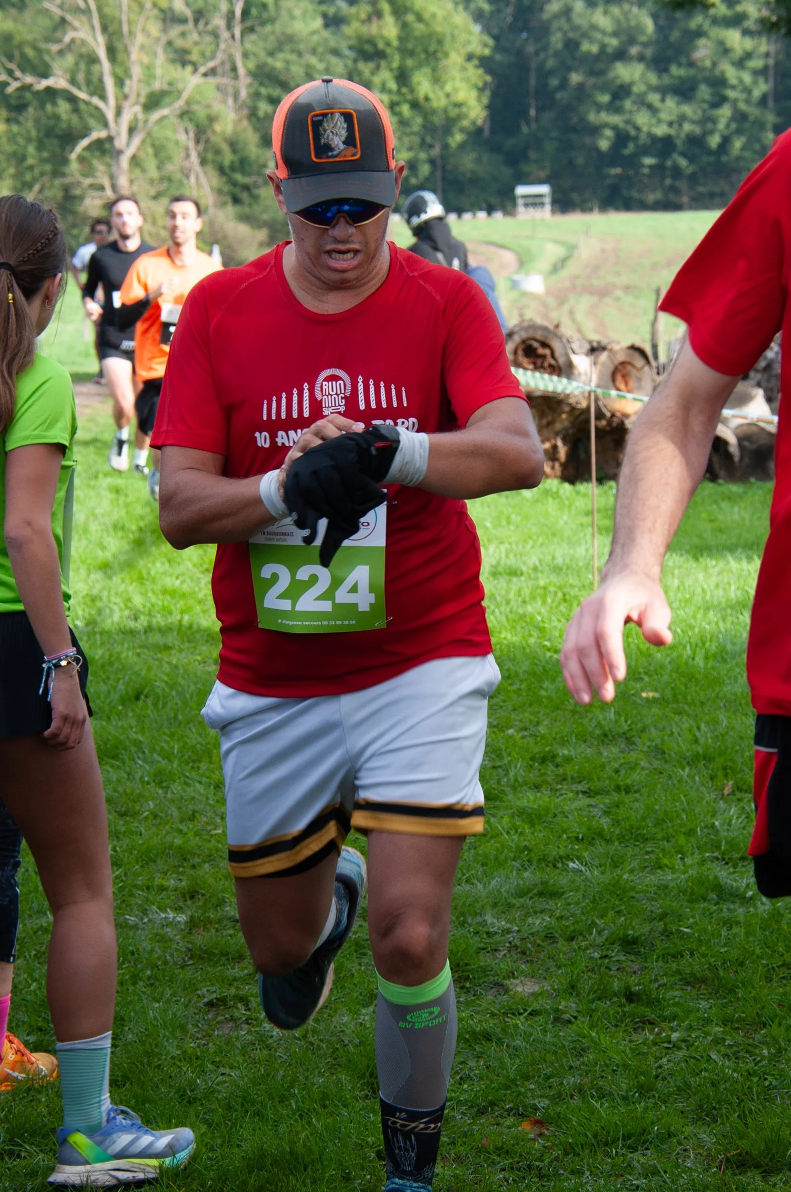 Un homme court lors d'une course en plein air, portant un dossard avec le numéro 224, une casquette, des lunettes de soleil, un t-shirt rouge, des shorts blanc avec des rayures noires et jaunes, et des chaussettes de compression grises. Il semble êtr