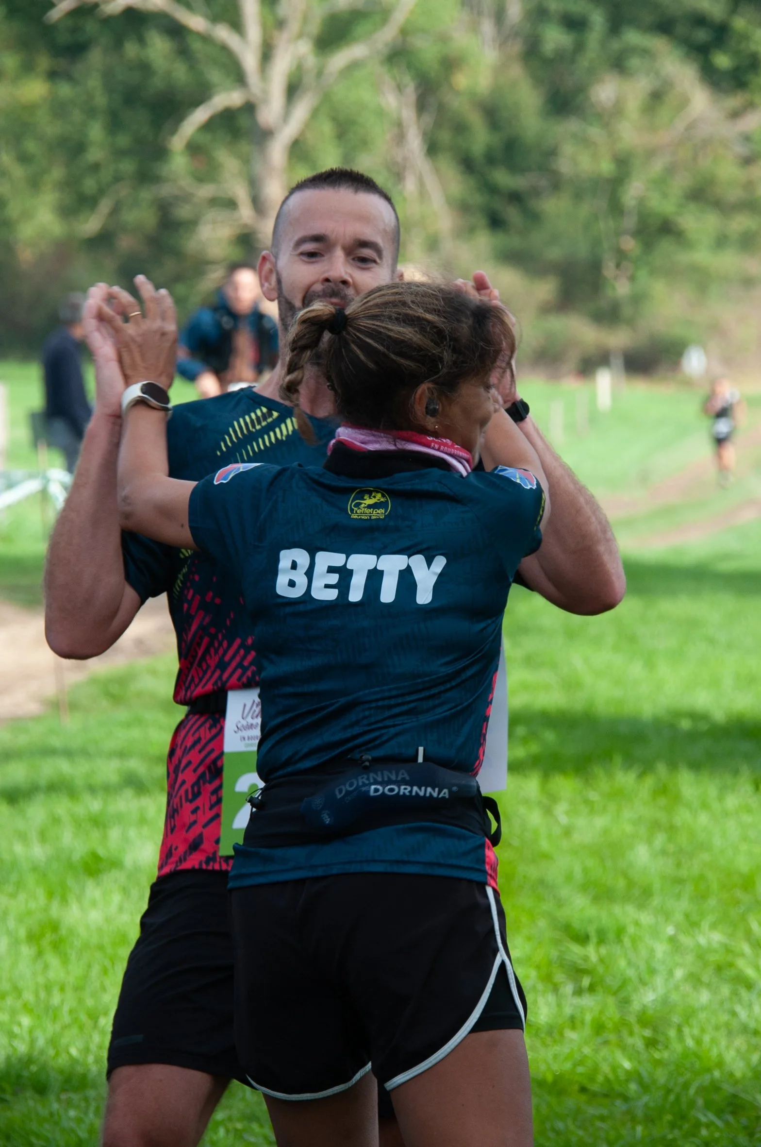 Deux coureurs se félicitent après une course en plein air, avec un fond verdoyant et quelques autres participants à l'arrière.