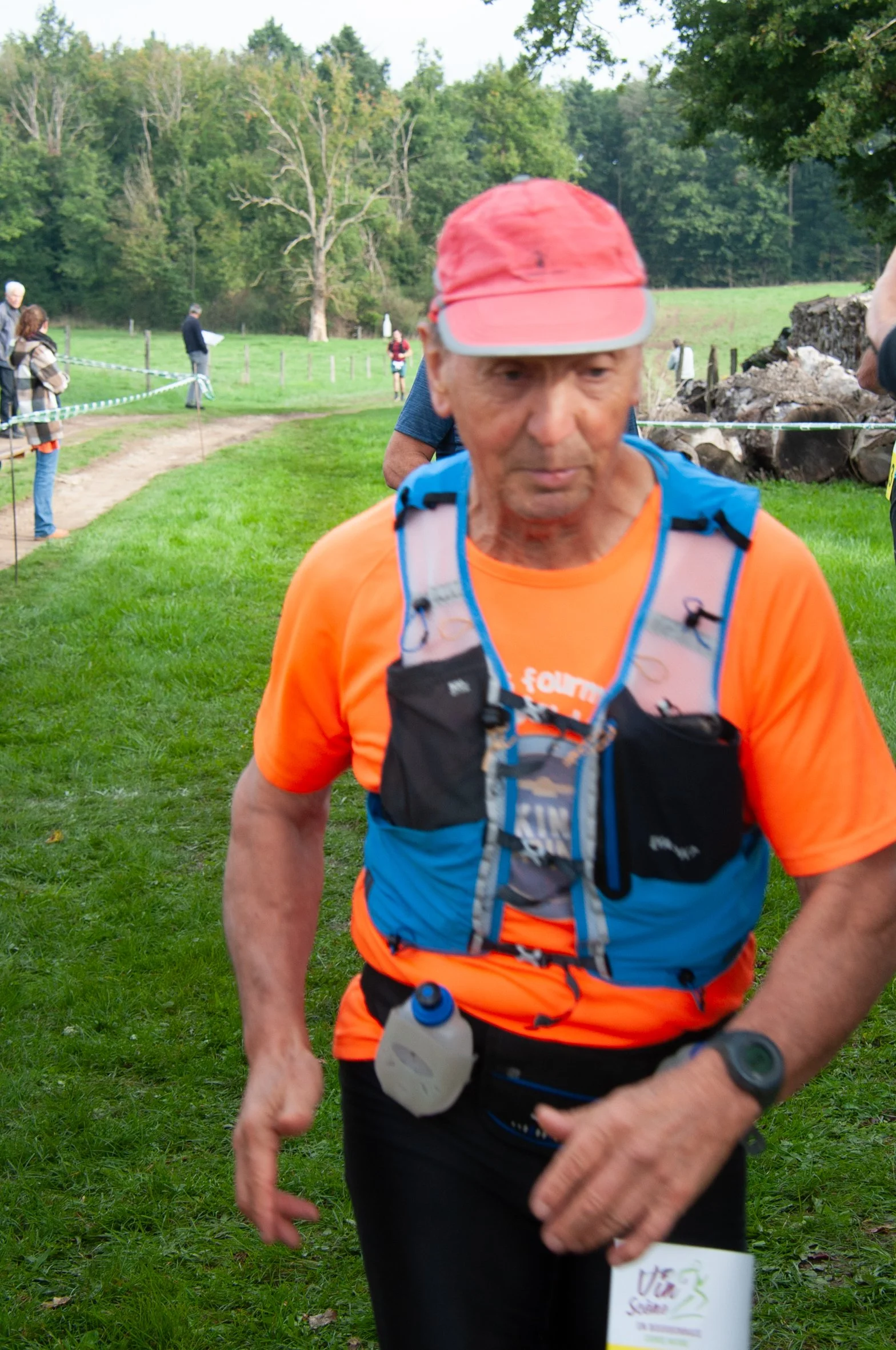 Un homme âgé participant à une course en plein air, portant une casquette rouge, un t-shirt orange, un gilet de randonnée bleu, et une montre, dans un environnement naturel avec de l'herbe verte et des arbres en arrière-plan.