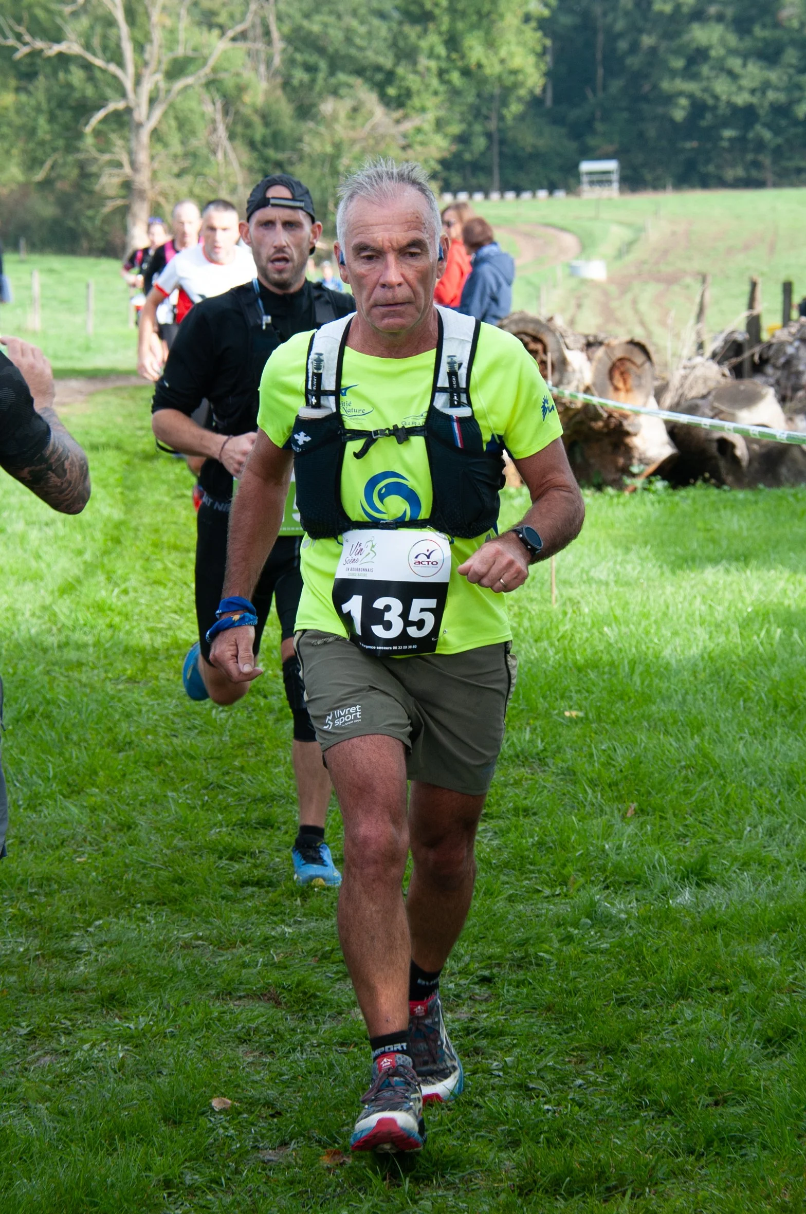 Un homme âgé portant un maillot jaune fluo, un numéro 135, participe à une course en plein air avec d'autres coureurs derrière lui, dans un environnement naturel avec de l'herbe verte et des arbres.
