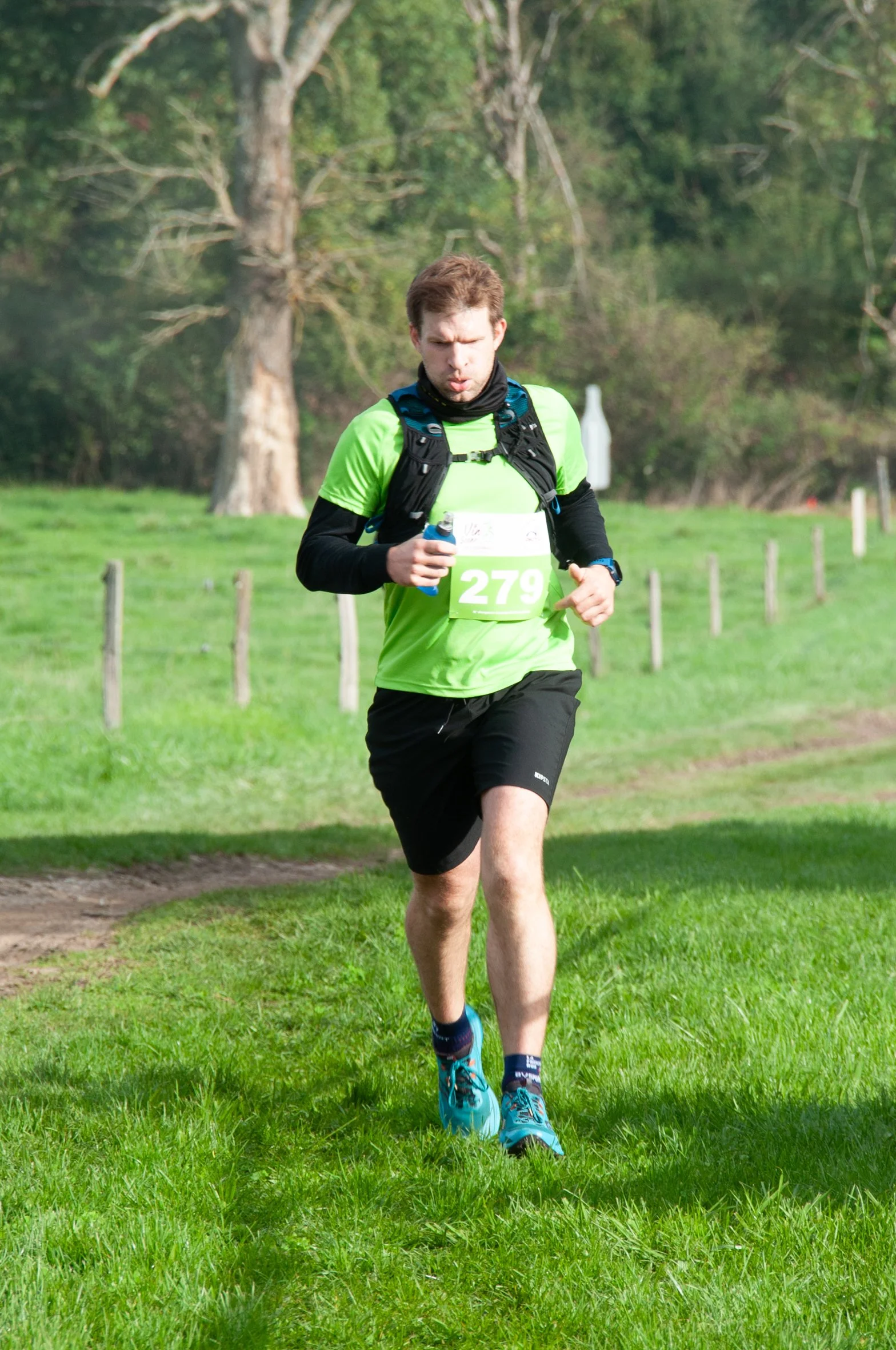 Un homme court dans un parc avec un t-shirt vert, un short noir, des chaussures de course bleues, portant un dossard numéro 279, en pleine course à pied dans un environnement vert avec des arbres et une pelouse.