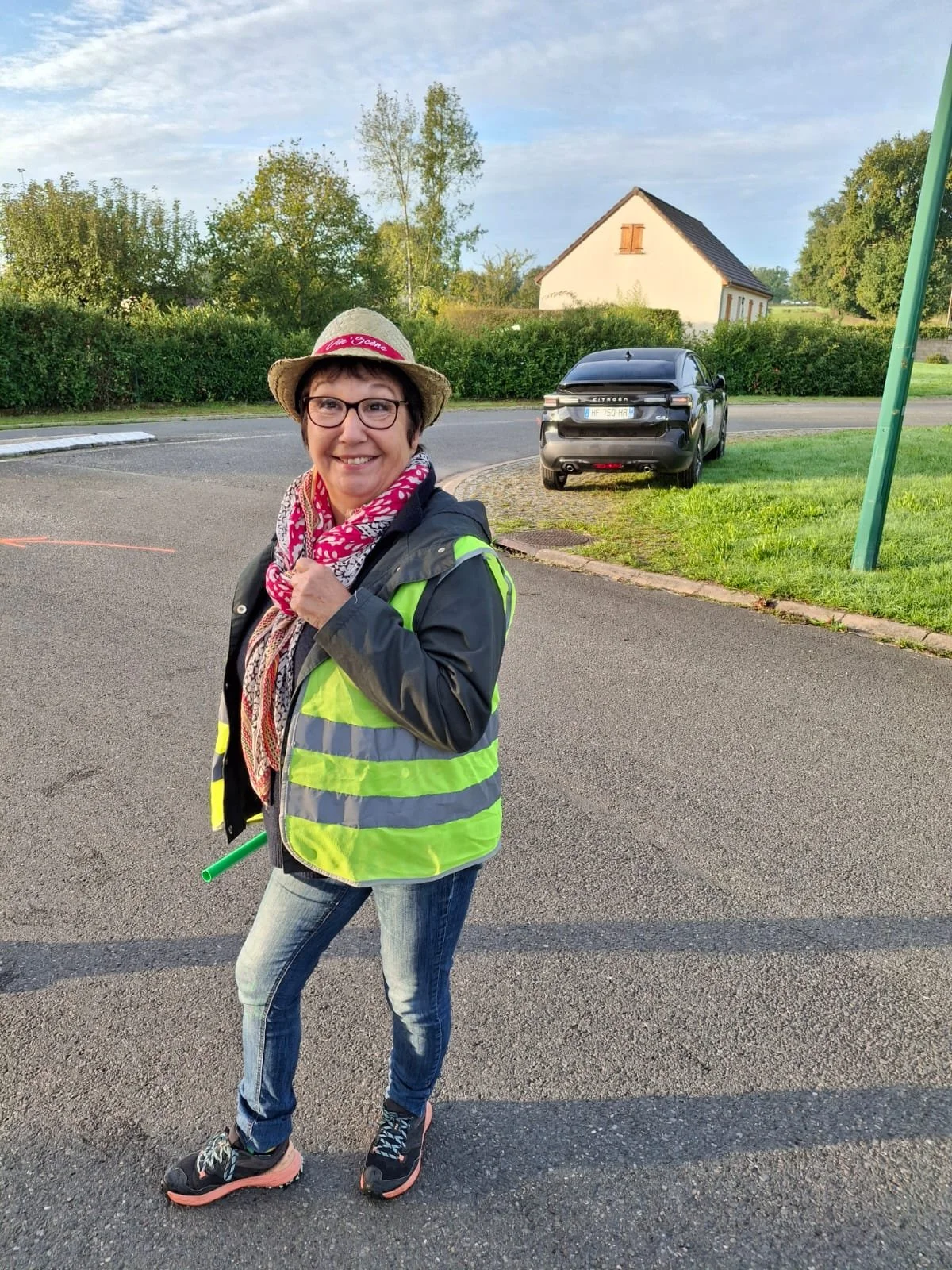 Femme souriante portant un gilet jaune réfléchissant, un chapeau en paille avec un ruban rose, des lunettes, un foulard rouge et blanc, debout sur le bord d'une route avec une voiture noire en arrière-plan, en soirée.