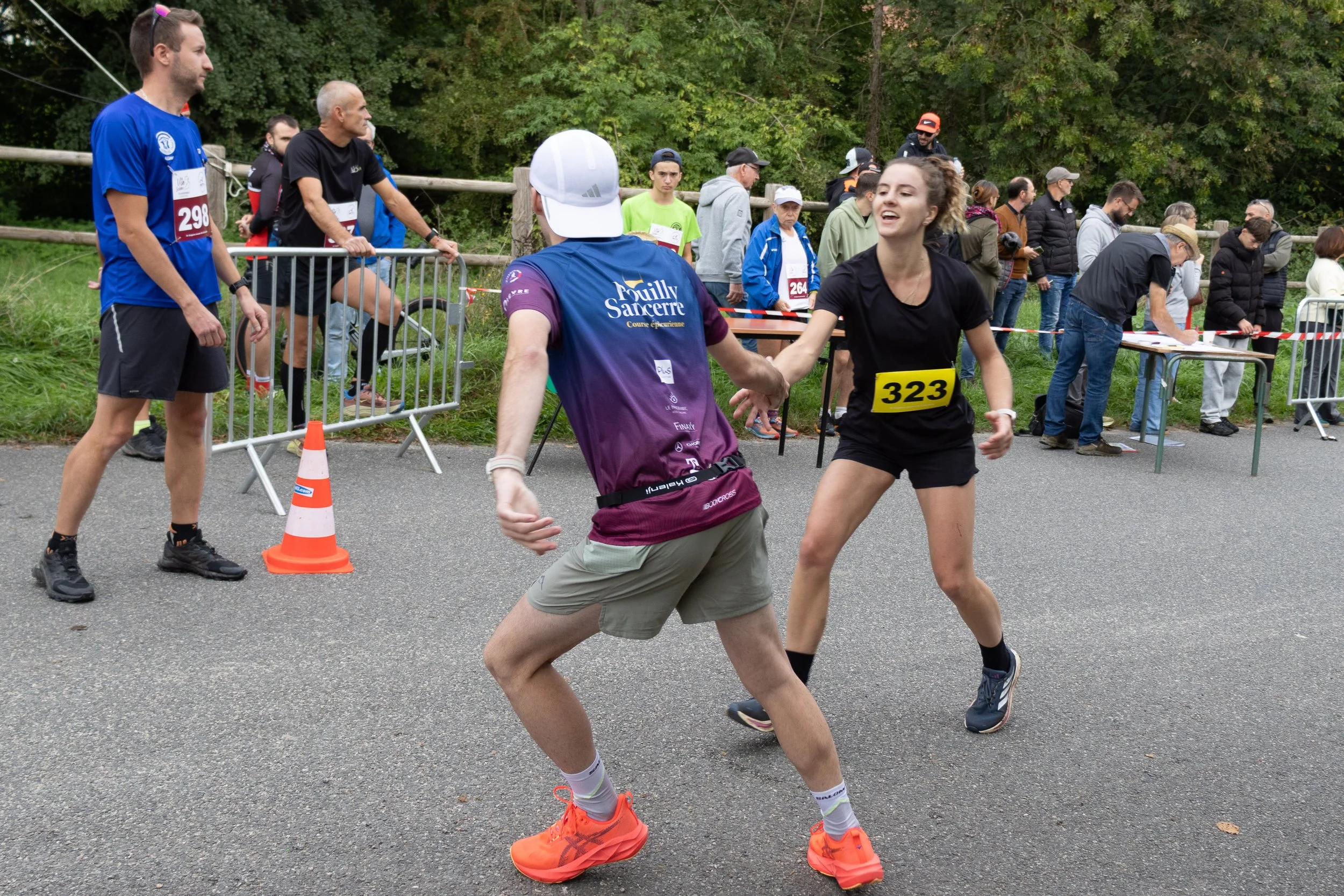 Deux coureurs souriants se donnent une poignée de main à la fin d'une course, entourés par d'autres participants et spectateurs, dans un environnement extérieur avec des arbres.