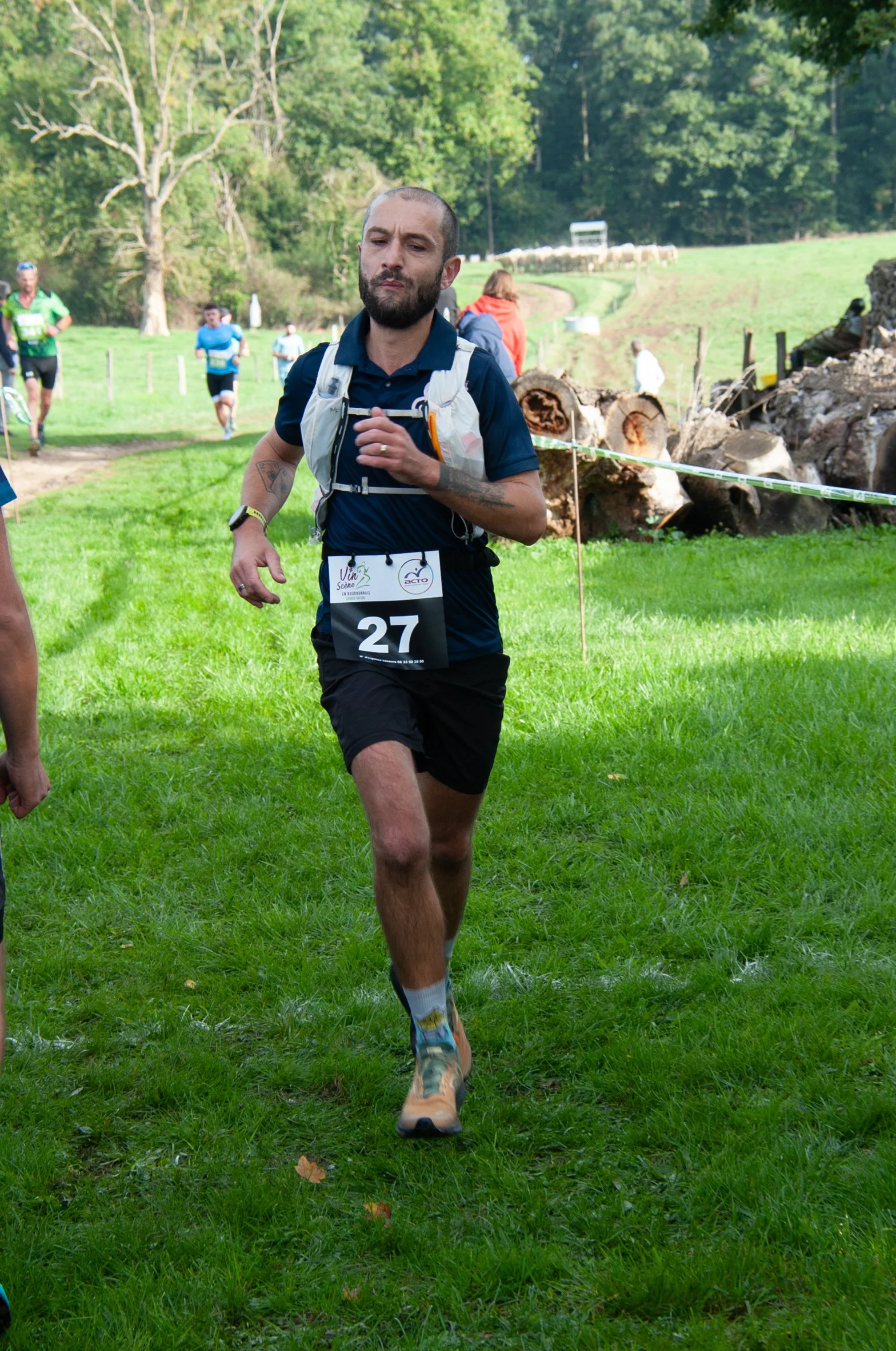 Un homme court lors d'une course en plein air, portant un dossard numéro 27, habillé d'un t-shirt bleu, d'un short noir, et d'une ceinture d'hydratation, avec un décor de forêt et de champs en arrière-plan.