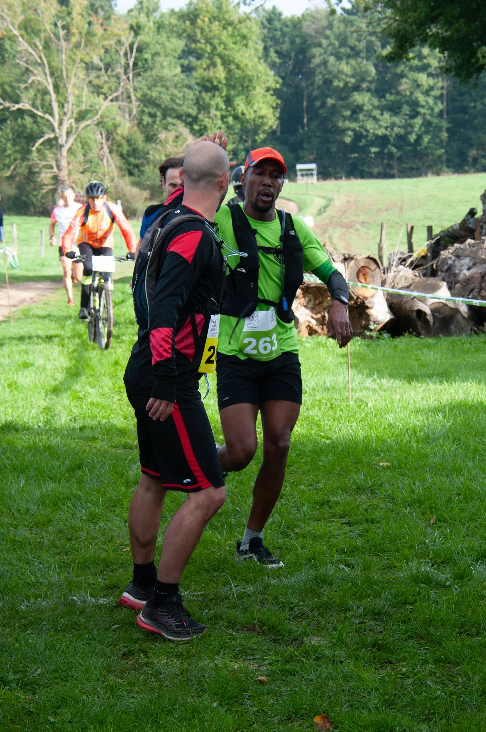 Deux hommes discutent lors d'une course ou randonnée en plein air, avec des cyclistes en arrière-plan, sur un chemin herbeux entouré d'arbres.