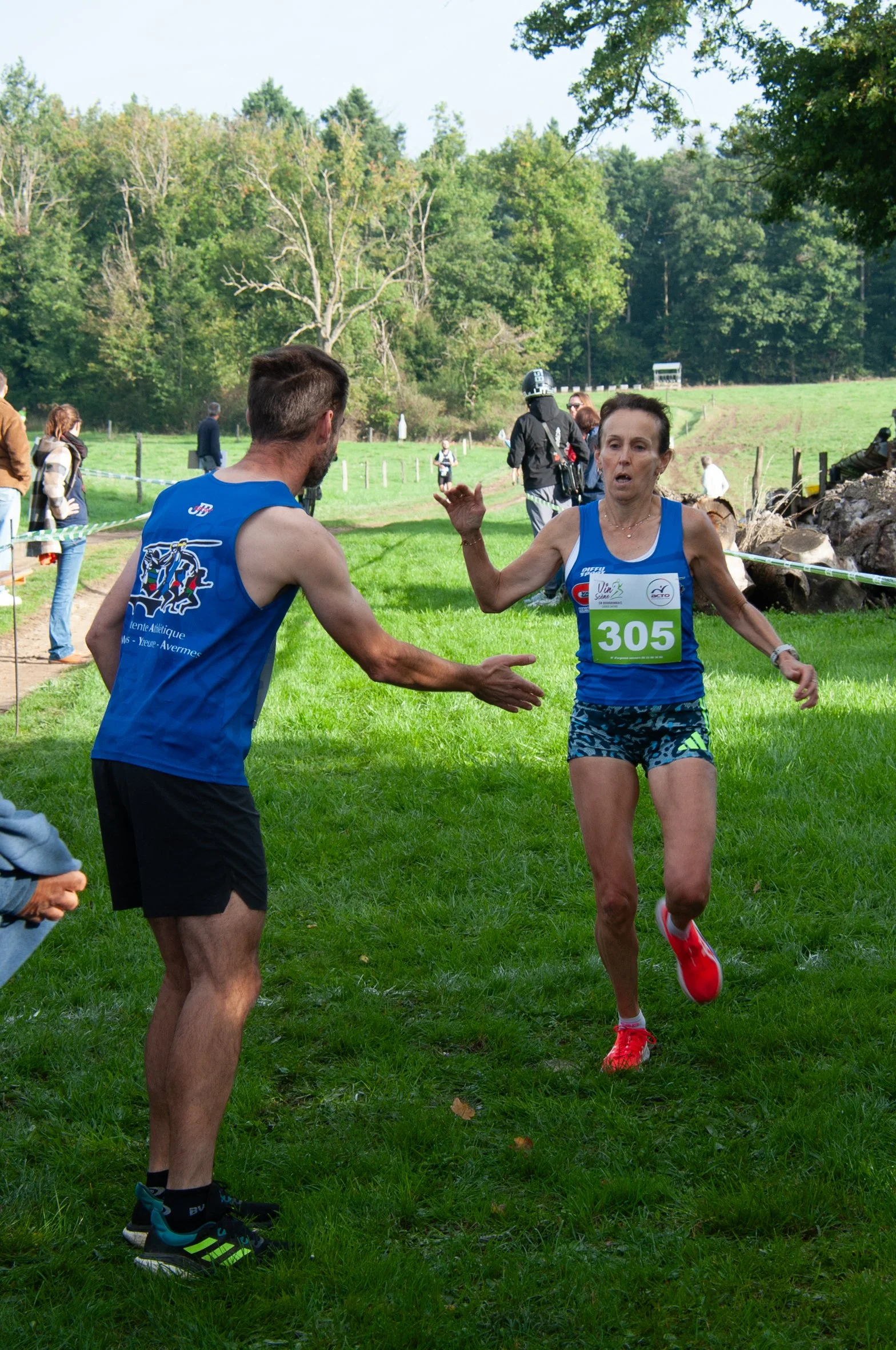 Un coureur à la course, portant un dossard vert avec le numéro 305, est en train de recevoir un encouragement ou une aide d'une personne en veston bleu lors d'une course en plein air dans un parc avec des arbres.