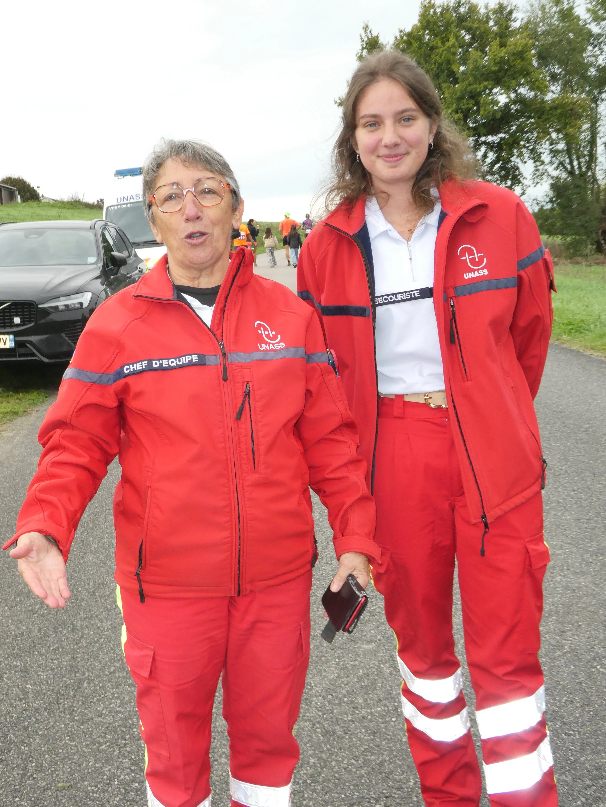 Deux femmes portent des uniformes rouges avec des inscriptions UNASS et sont en extérieur, avec des voitures et des arbres en arrière-plan.