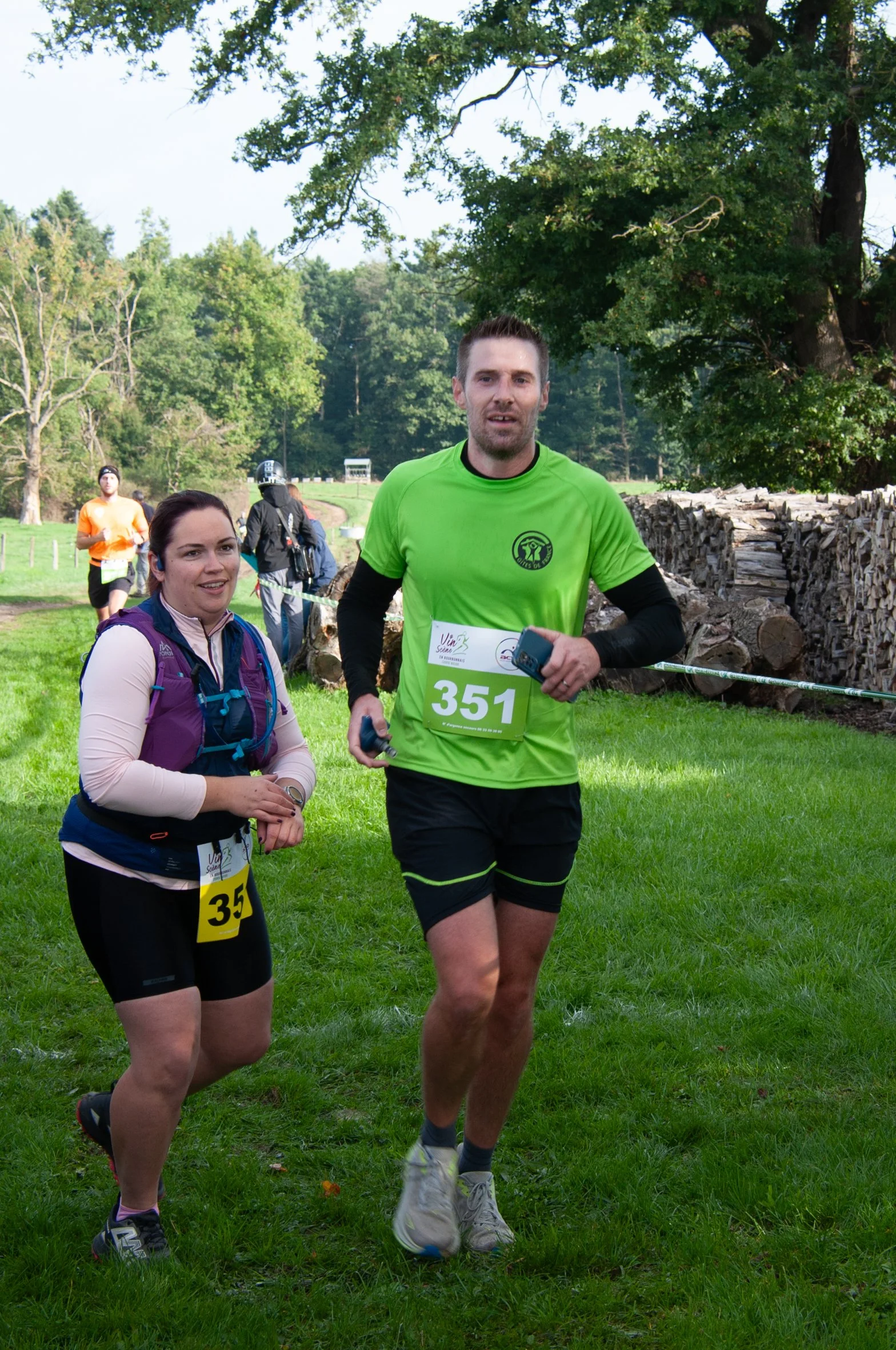 Deux coureurs participent à une course en extérieur, portant des numéros 351 et 35, avec un groupe d'autres participants en arrière-plan, dans un environnement verdoyant avec des arbres et une pile de bois.