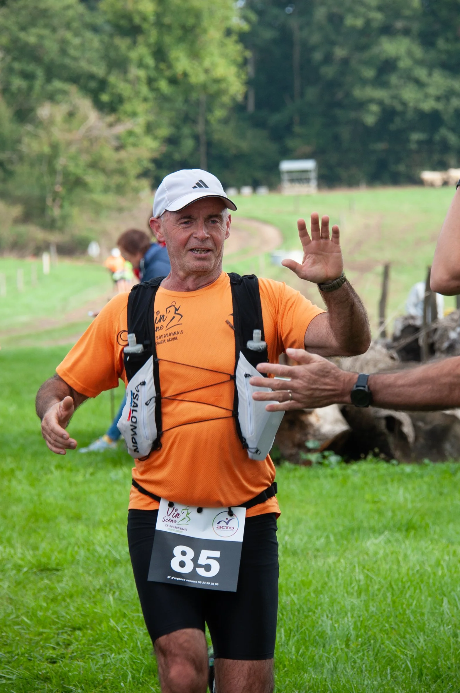 Un homme en tenue de course, portant un gilet d'hydratation et un numéro de course 85, participe à une compétition en plein air dans un environnement verdoyant, recevant un encouragement d'une autre personne.