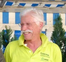 Un homme âgé avec des cheveux blancs, portant une chemise jaune vif, souriant, dans un environnement extérieur avec des décorations bleues et blanches et des plantes vertes.