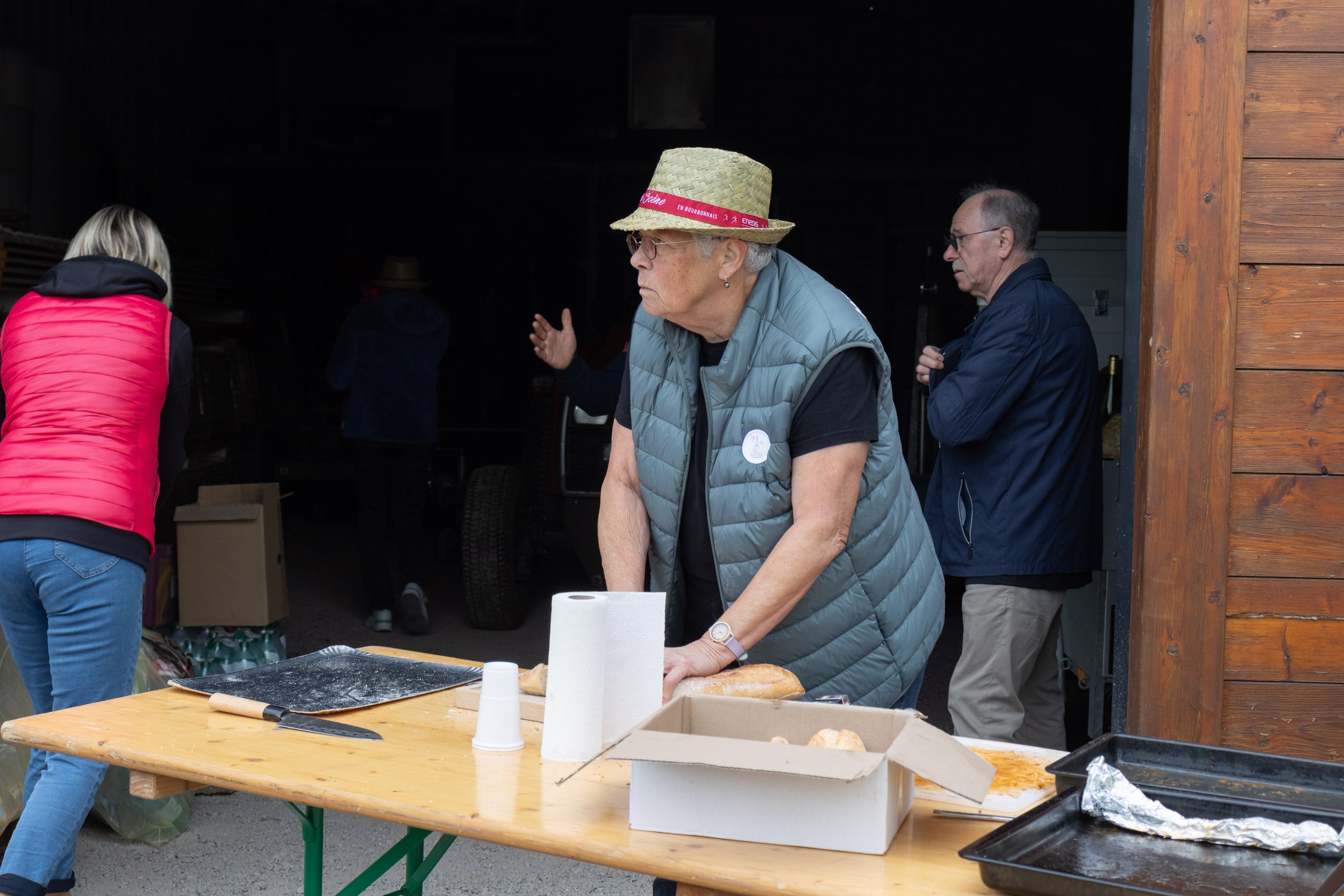Une femme âgée portant un chapeau en paille, une veste grise et un t-shirt noir, prépare du pain sur une table en bois. Autour, plusieurs personnes discutent ou préparent quelque chose, dans un espace intérieur sombre ou un garage avec un bâtiment en