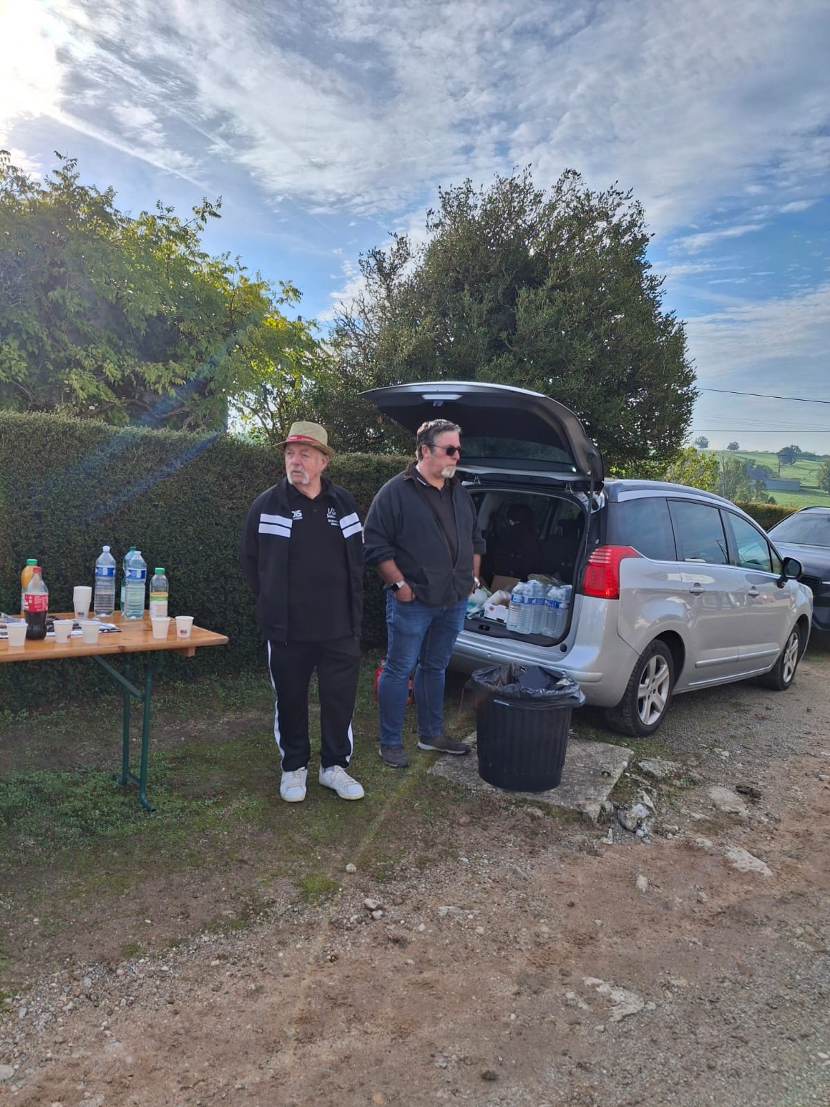 Deux hommes se tiennent devant une voiture avec le coffre ouvert, à côté d'une table avec des bouteilles et des gobelets, dans un environnement extérieur avec des arbres et un ciel en partie nuageux.