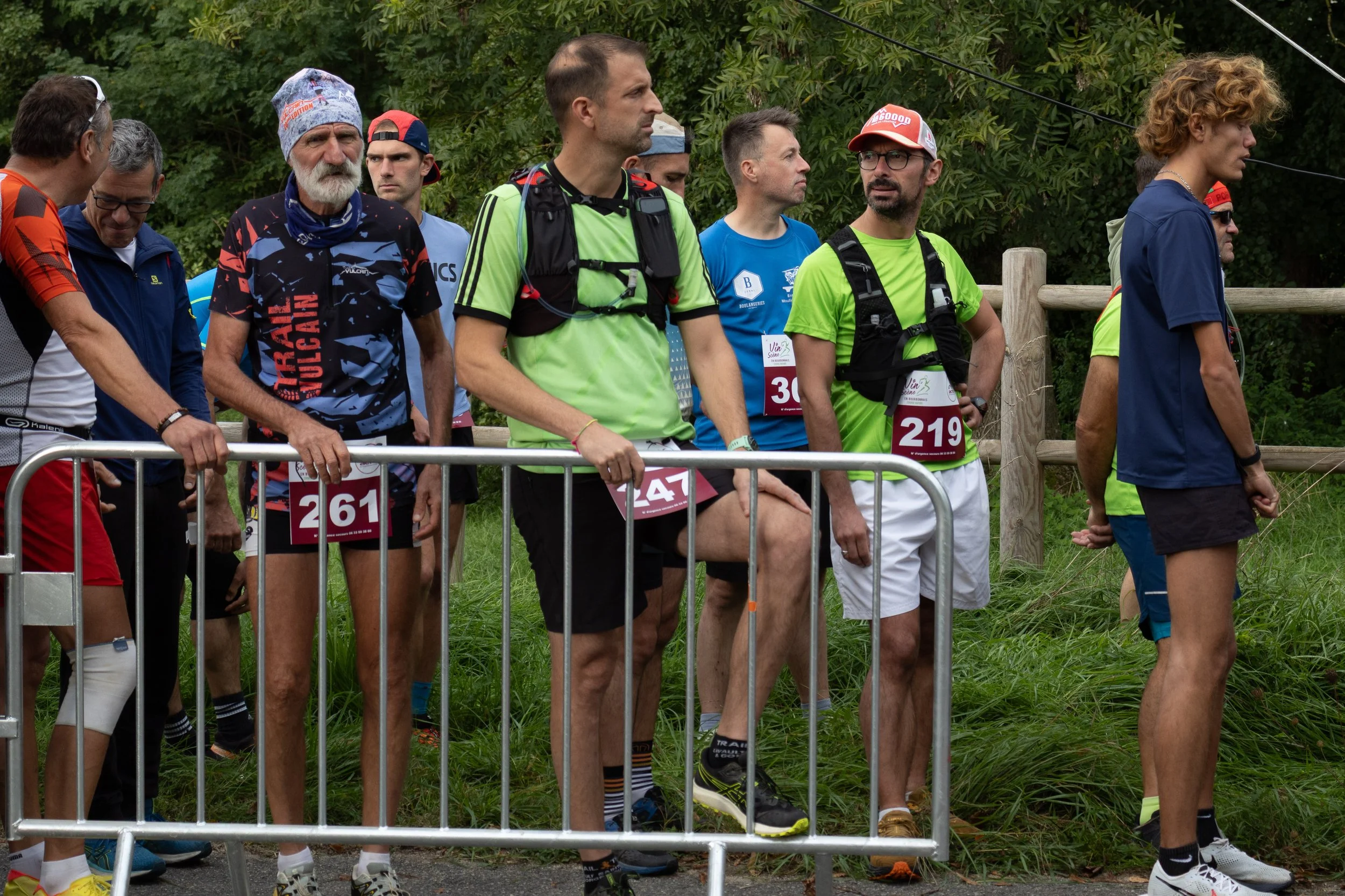 Groupe de coureurs en attente au début d'une course, portant des numéros de dossard, dans un environnement extérieur avec de la verdure.