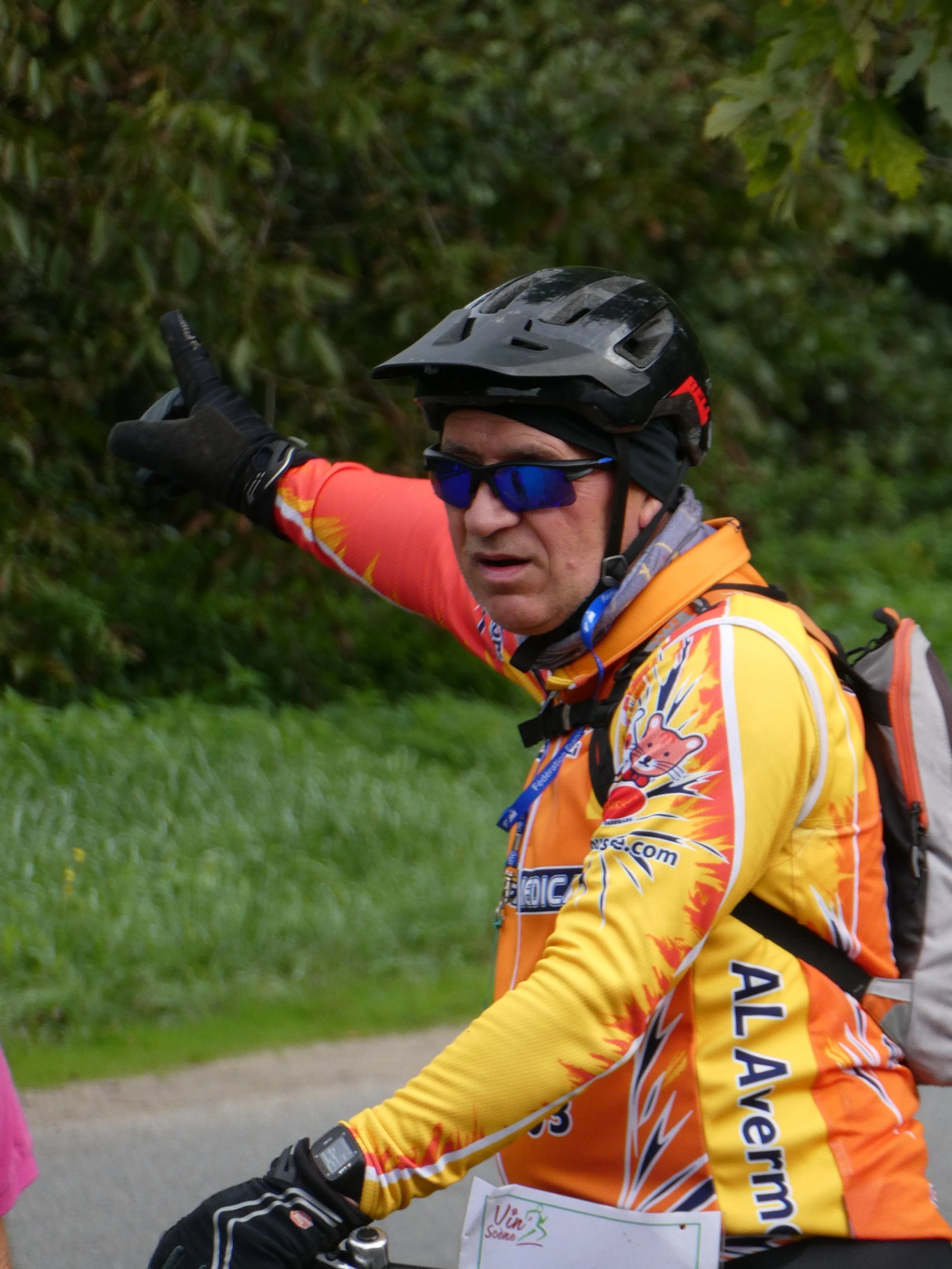 Un homme portant un casque de vélo, des lunettes de soleil, un maillot de cyclisme orange et jaune avec des motifs de lion, un sac à dos, et un ordinateur de sport, indique vers la gauche dans un environnement naturel.