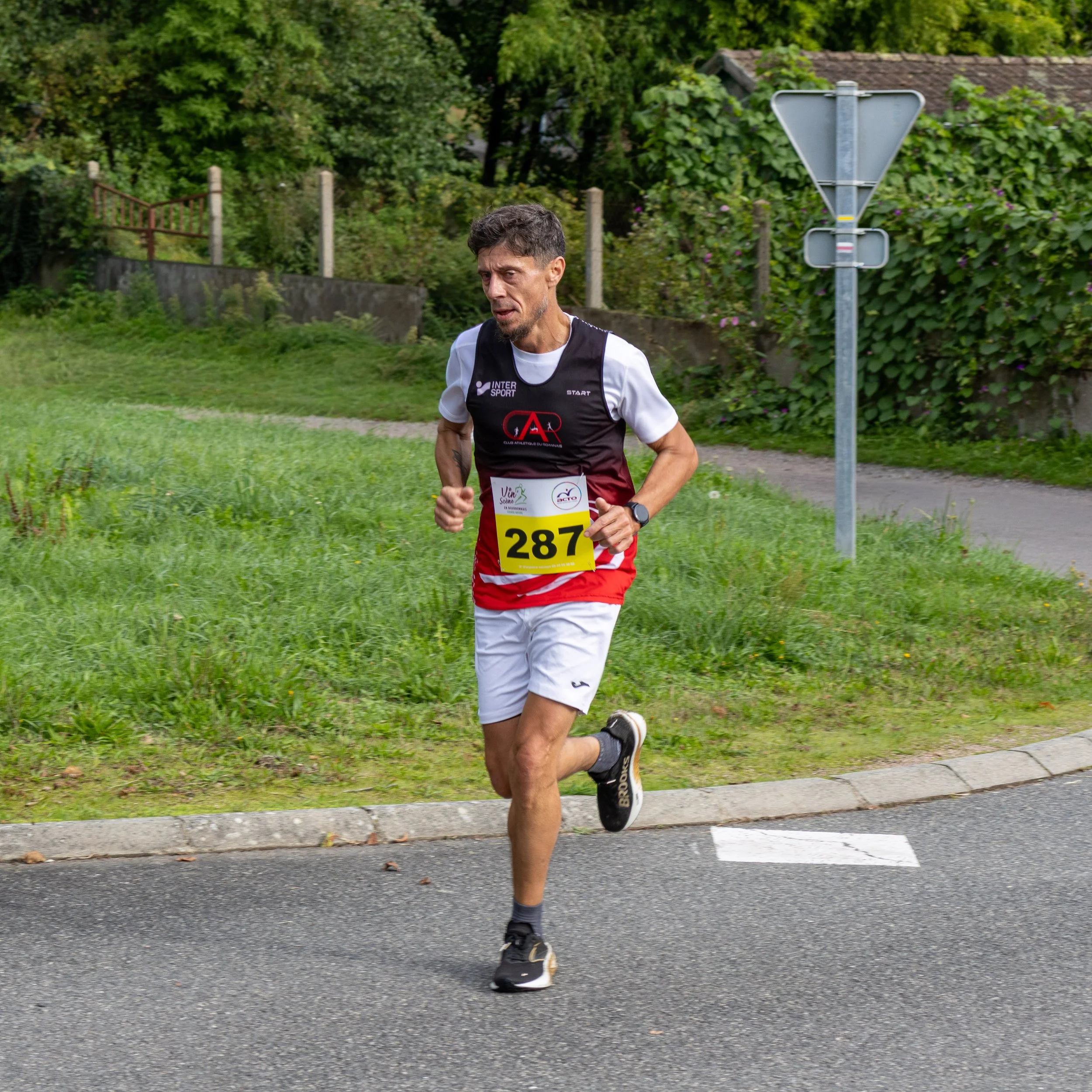 Un homme courant lors d'une compétition de course, portant un maillot sans manches noir et blanc avec un dossard numéro 287, dans un environnement extérieur verdoyant.