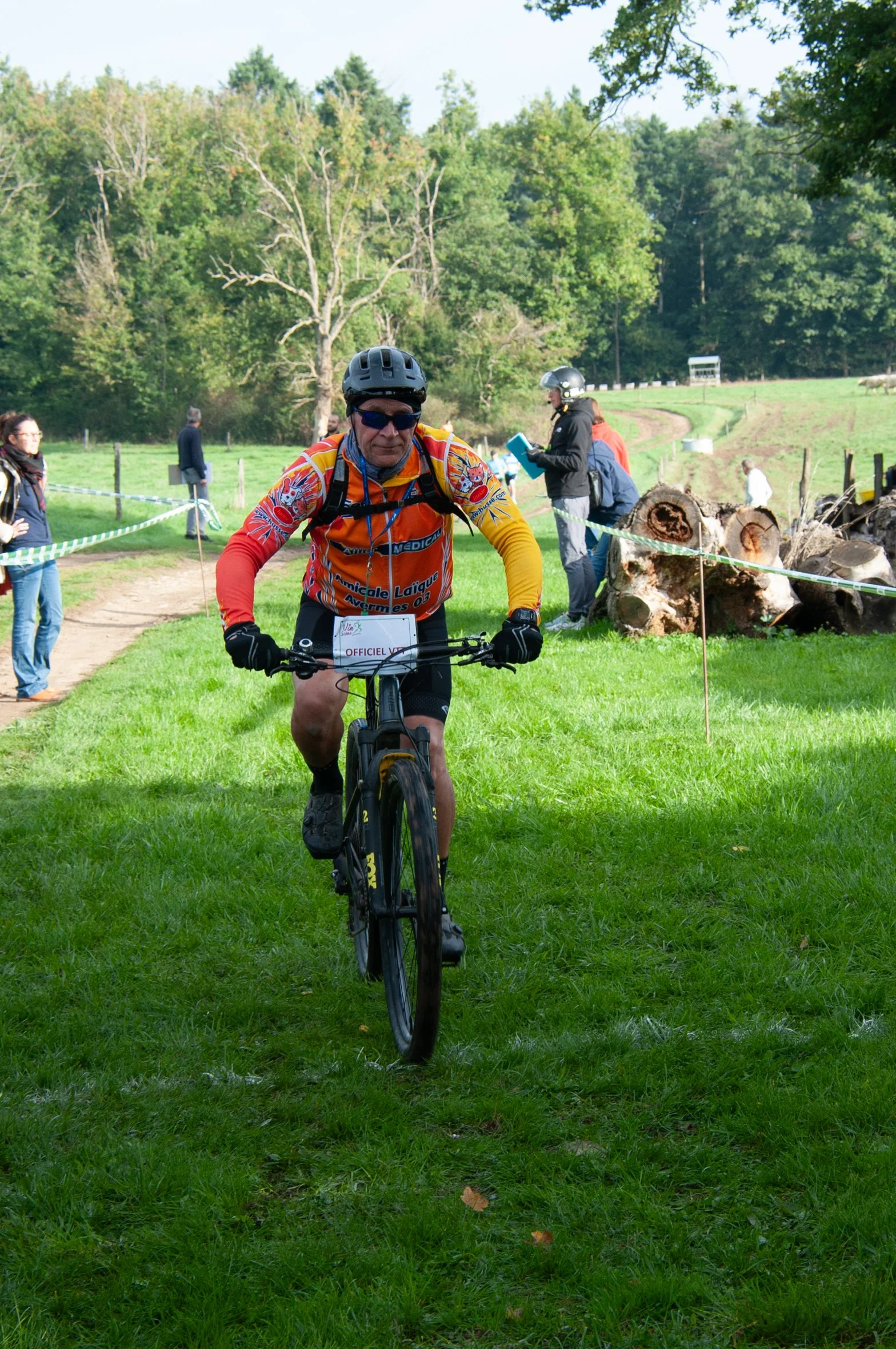 Un homme en vélo, portant un casque et des lunettes, participe à une course en plein air dans un espace verdoyant. Il porte un maillot orange avec des motifs colorés et un numéro de course fixé à son vélo. Quelques spectateurs et organisateurs sont v