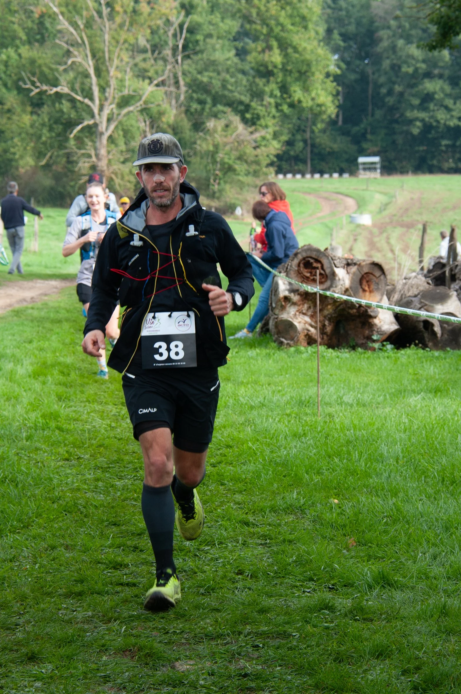 Un coureur participe à une course en plein air sur un sentier vert, portant un dossard numéro 38, avec d'autres personnes en arrière-plan.