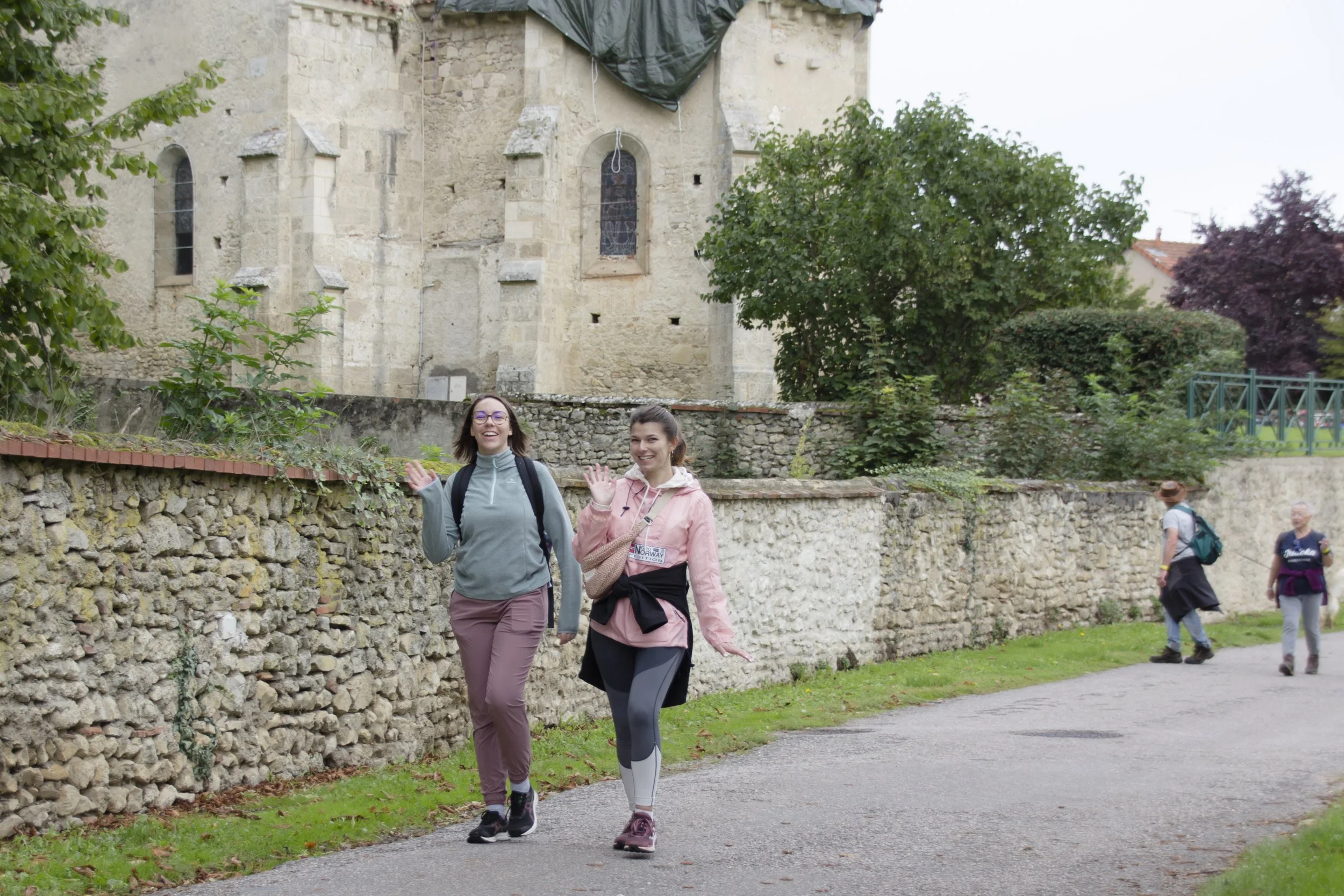 Deux femmes marchent et sourient en saluant, précisées par des sacs à dos, avec un vieux mur en pierre et un bâtiment en pierre ancienne en arrière-plan, dans un environnement urbain ou historique.