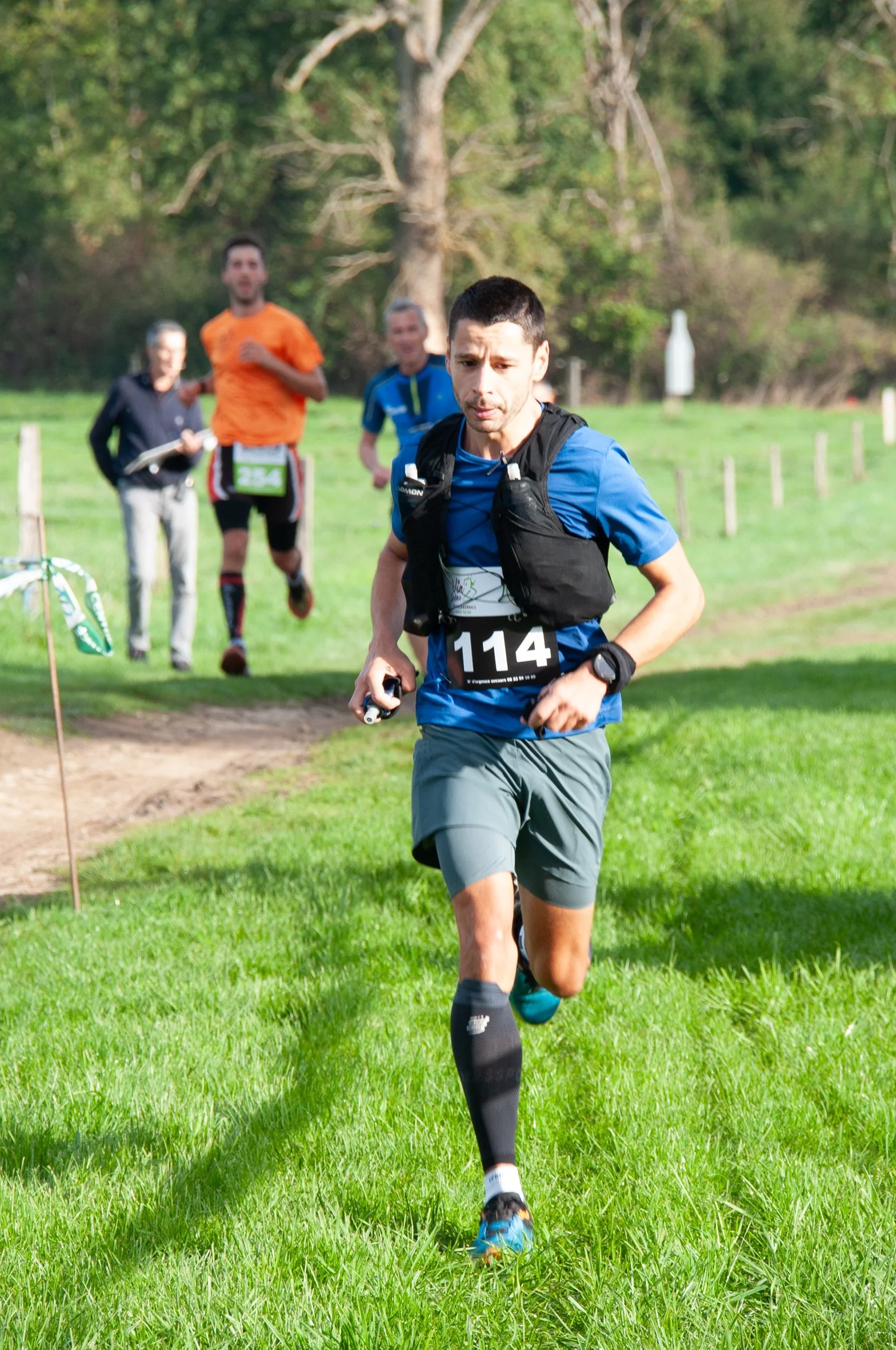 Un homme en course à pied dans un parc lors d'une compétition, portant un maillot bleu, un bib numéro 114, et une veste noire sur les épaules, avec d'autres coureurs en arrière-plan.