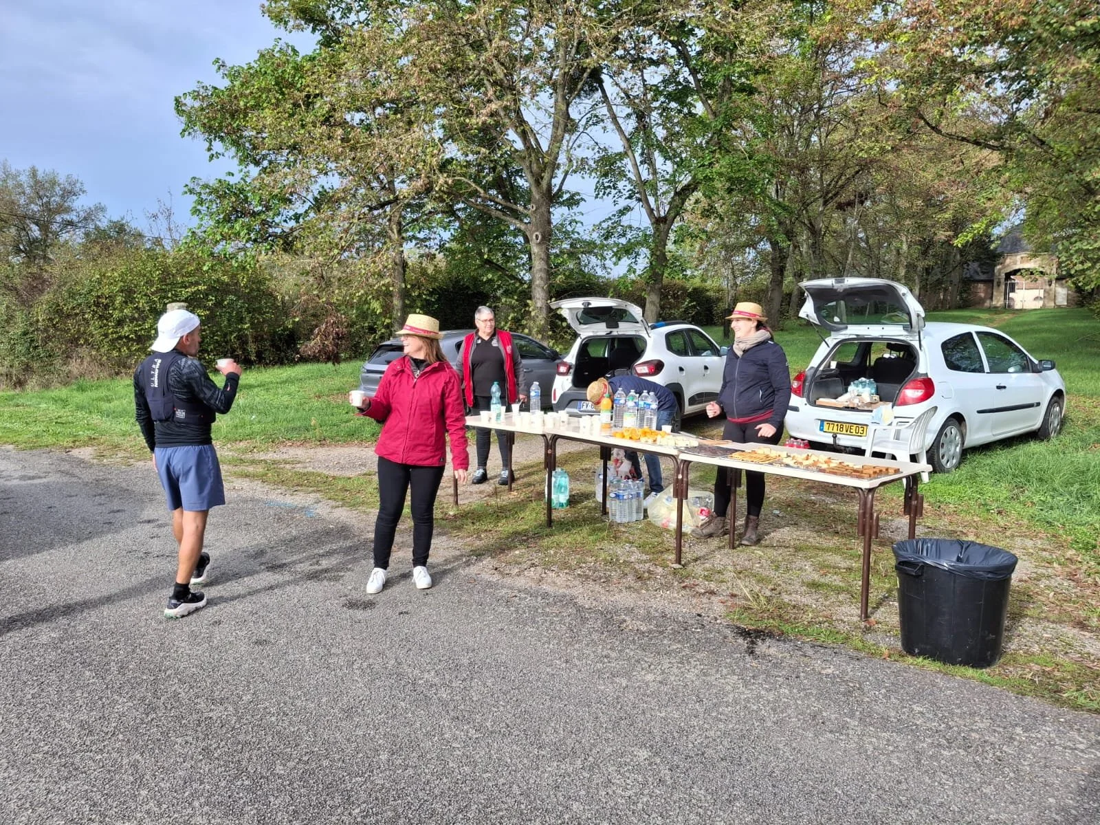 Regroupement de personnes près d'une table avec collations et boissons, en plein air, dans un parking entouré d'arbres.