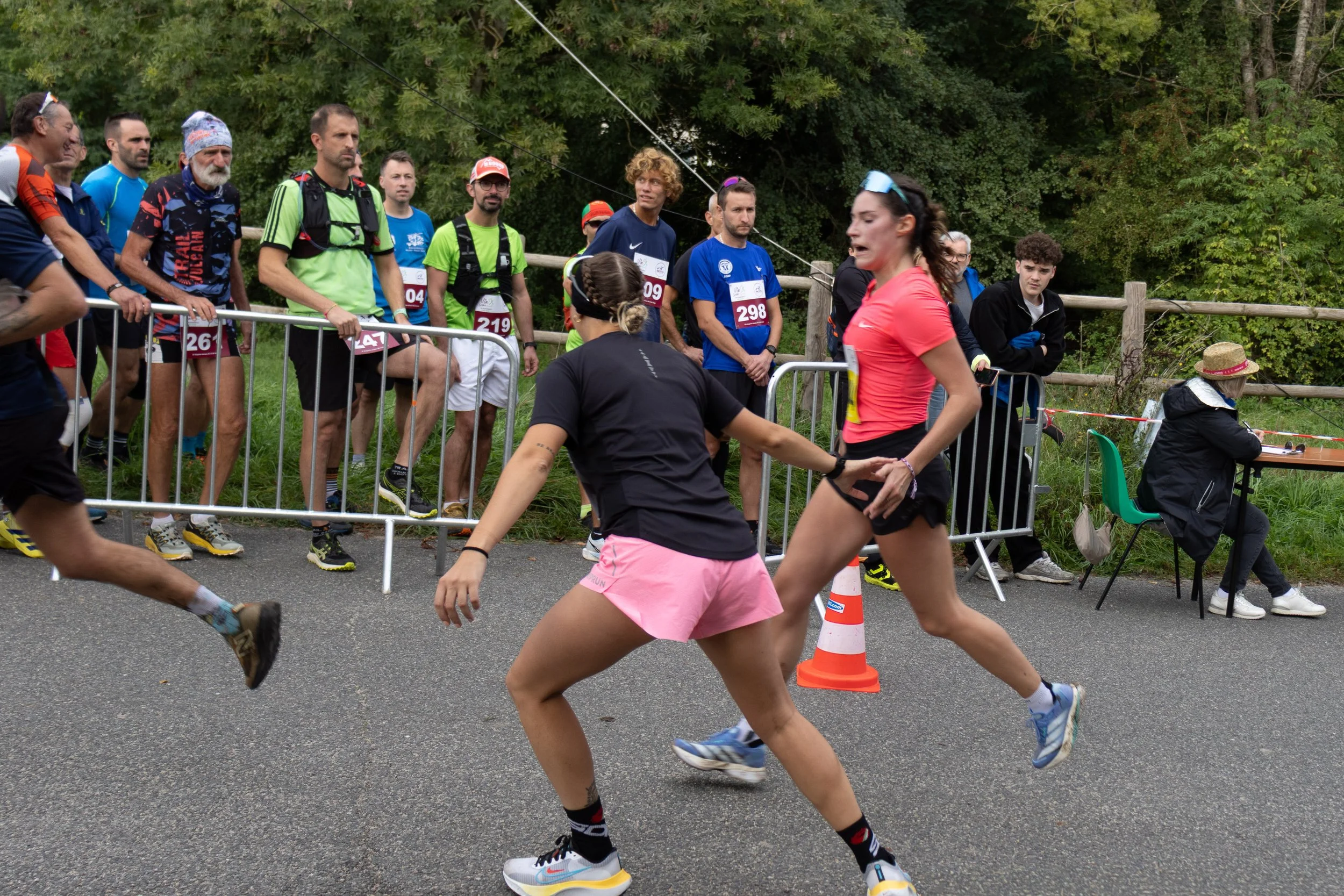 Deux femmes, avant ou après une course, semblent se disputer ou s'aider lors d'une compétition. En arrière-plan, plusieurs coureurs et spectateurs observant la scène, certains derrière une barrière en métal, dans un environnement extérieur avec des a